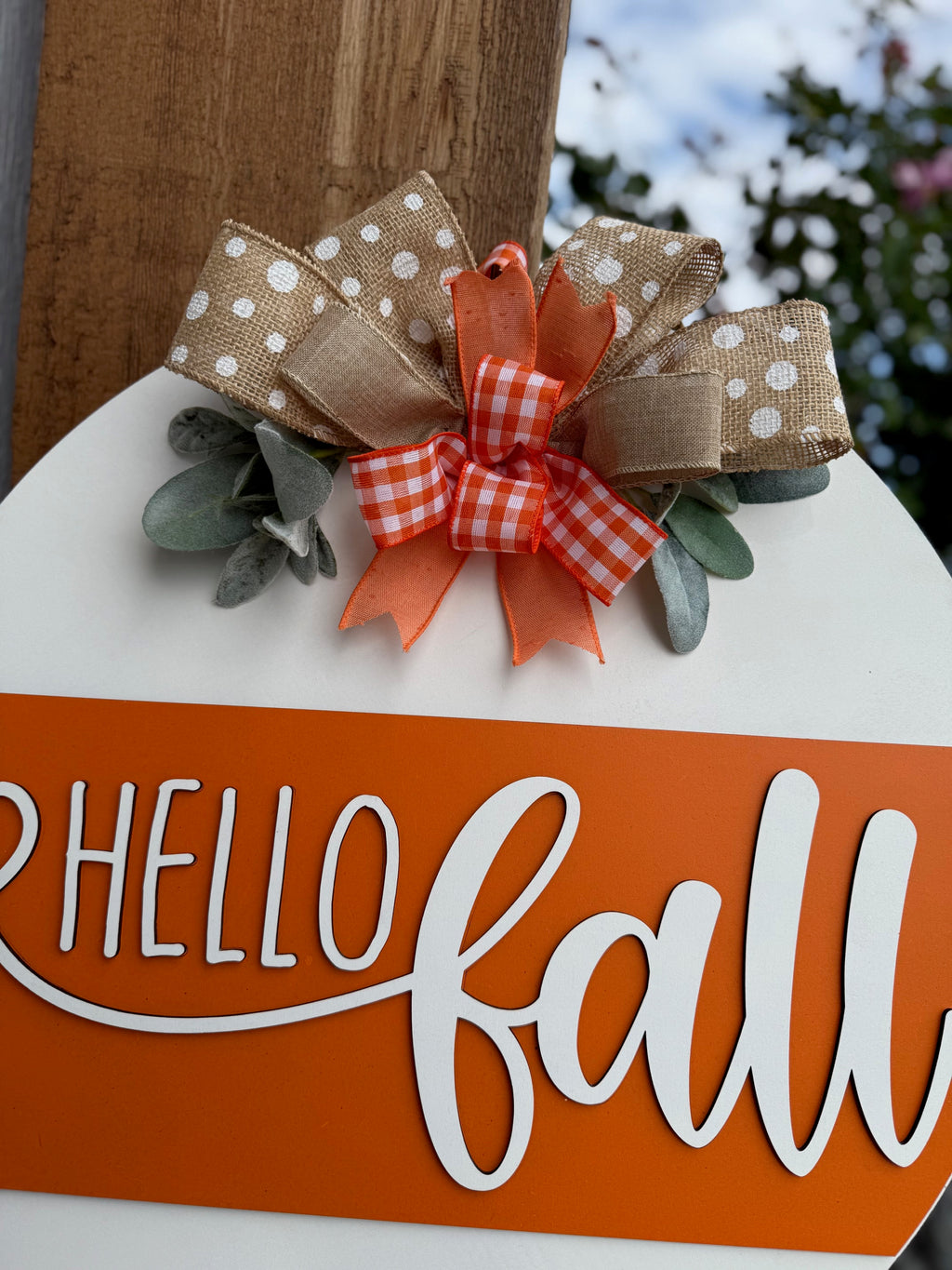 A round sign with Hello Fall in white script on an orange stripe, decorated with polka dot and checkered bows and faux greenery, is displayed on a white brick ledge near a garden.