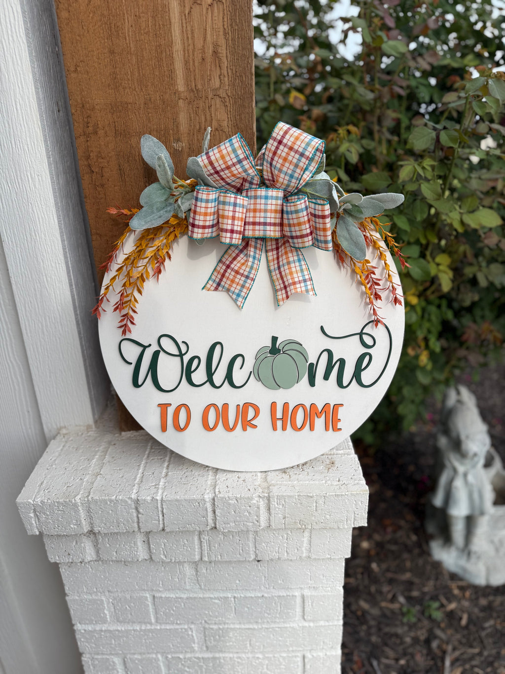 Round white sign with a plaid bow, greenery, and orange sprigs. It reads Welcome to our home, with a green pumpkin in place of the “o” in Welcome. The sign rests on a white brick ledge.