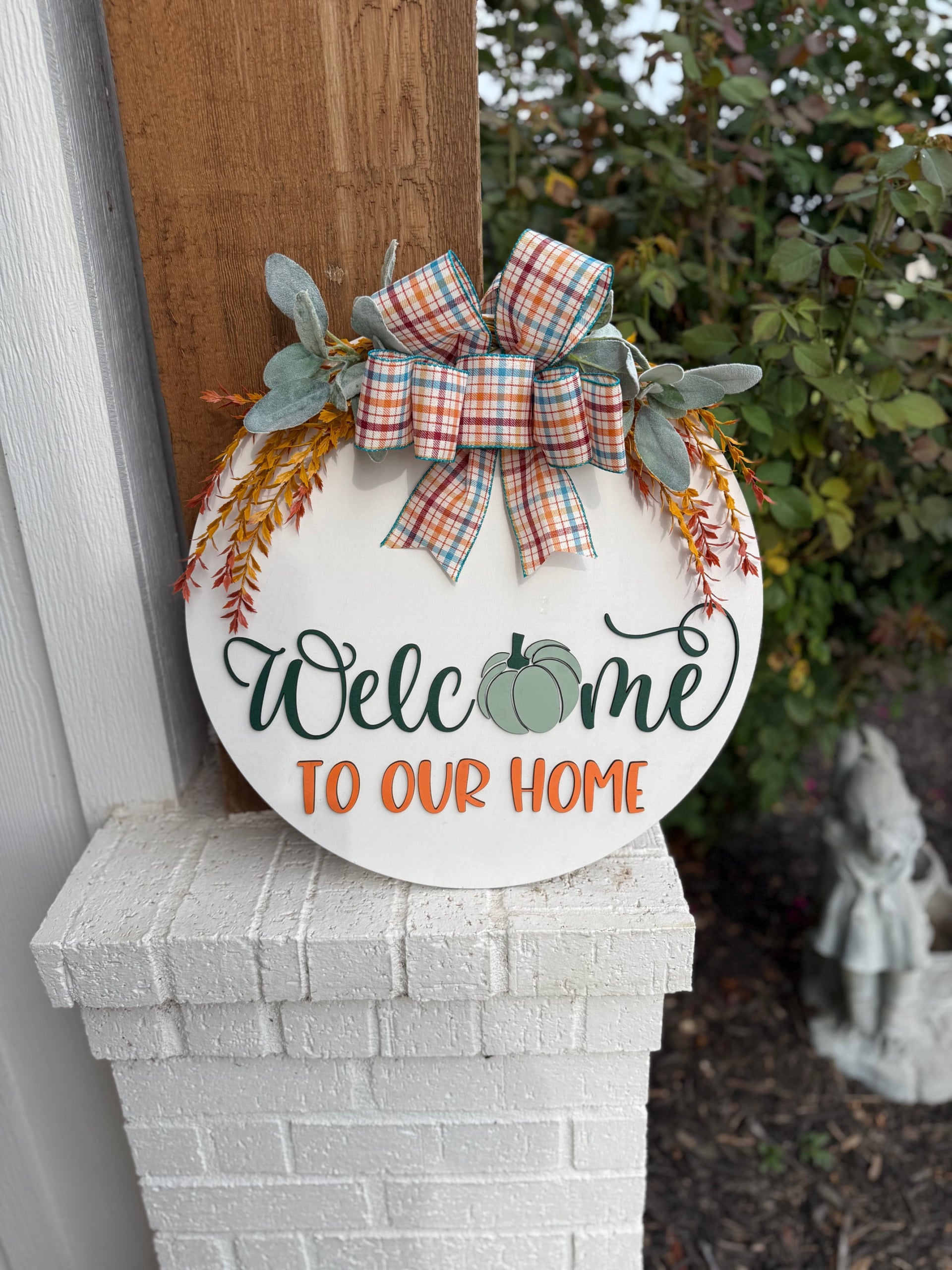 Round white sign with a plaid bow, greenery, and orange sprigs. It reads Welcome to our home, with a green pumpkin in place of the “o” in Welcome. The sign rests on a white brick ledge.
