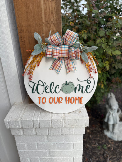 Round white sign with a plaid bow, greenery, and orange sprigs. It reads Welcome to our home, with a green pumpkin in place of the “o” in Welcome. The sign rests on a white brick ledge.