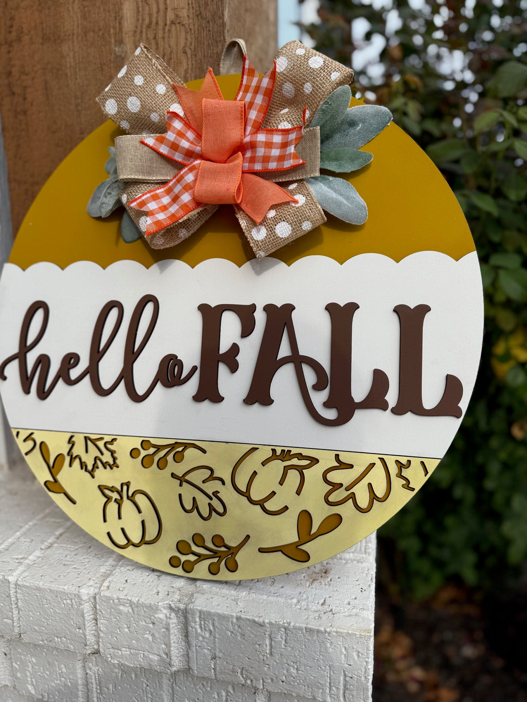 A round yellow and white sign with hello FALL written on it, decorated with a polka dot and plaid bow, autumn leaves, and placed on a white brick ledge next to a wooden post and some greenery.