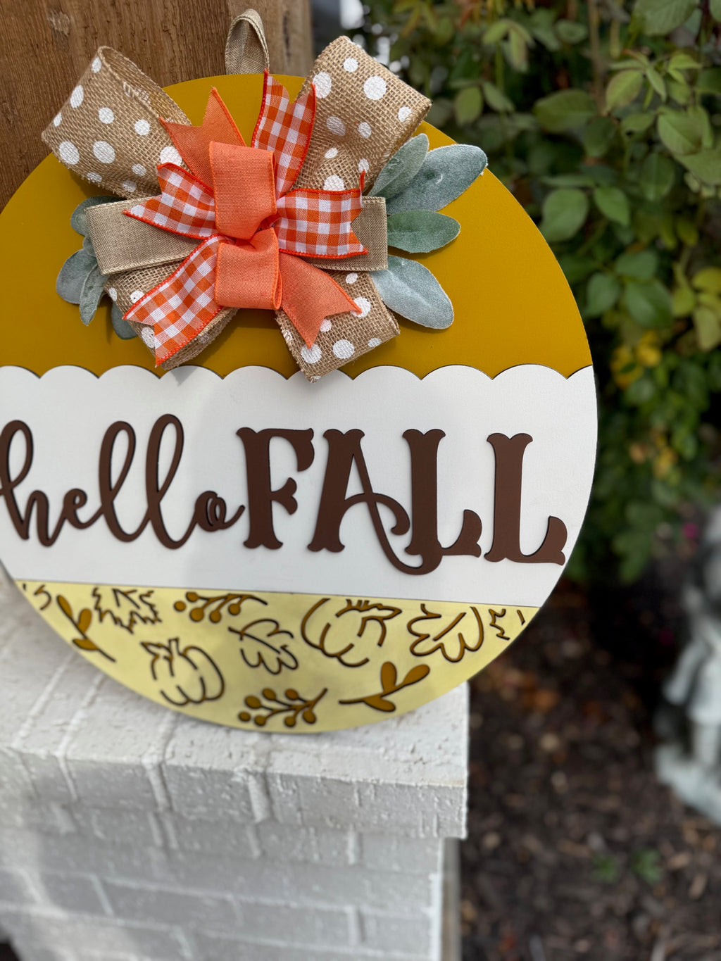 A round yellow and white sign with hello FALL written on it, decorated with a polka dot and plaid bow, autumn leaves, and placed on a white brick ledge next to a wooden post and some greenery.