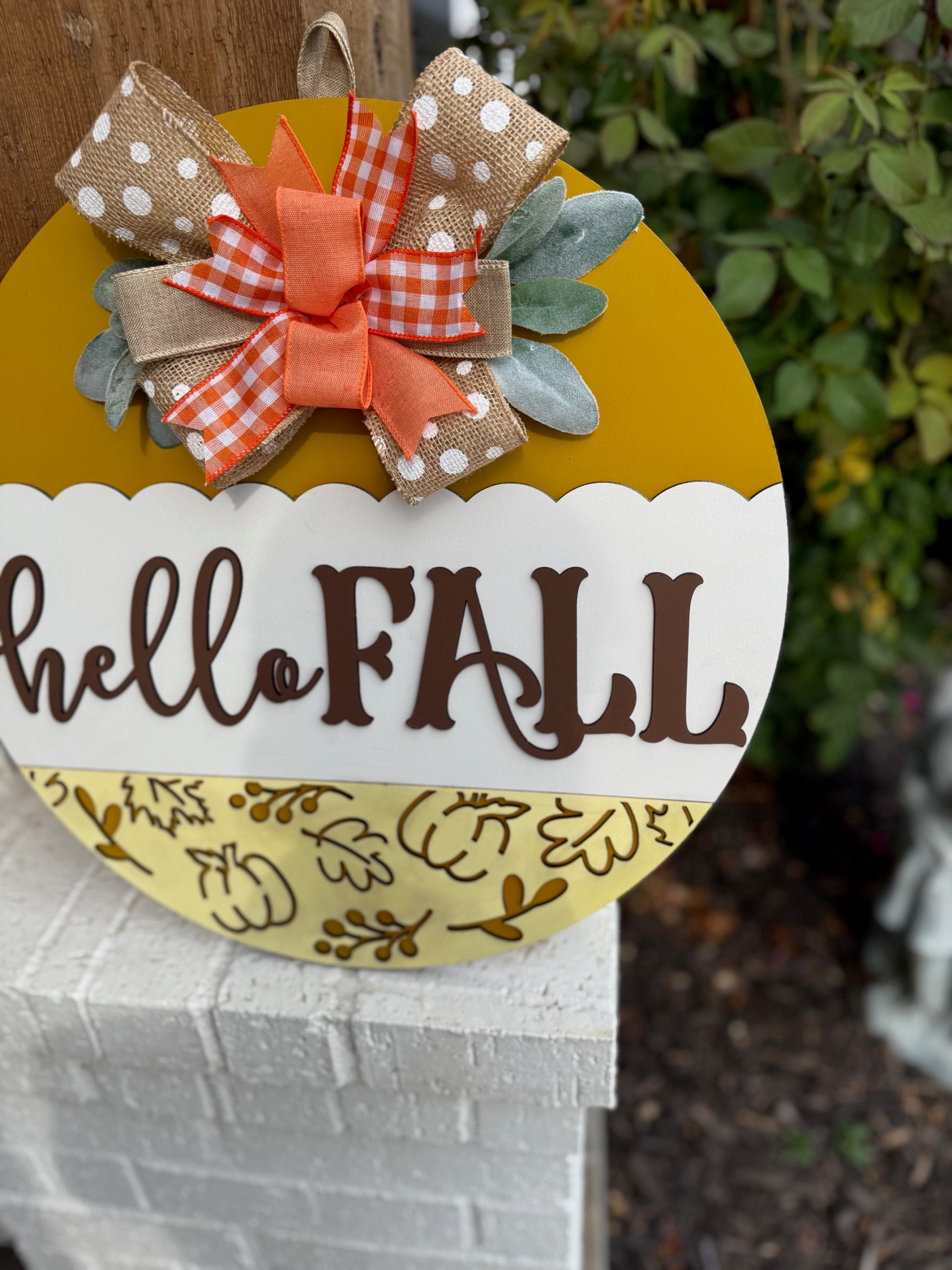 A round yellow and white sign with hello FALL written on it, decorated with a polka dot and plaid bow, autumn leaves, and placed on a white brick ledge next to a wooden post and some greenery.