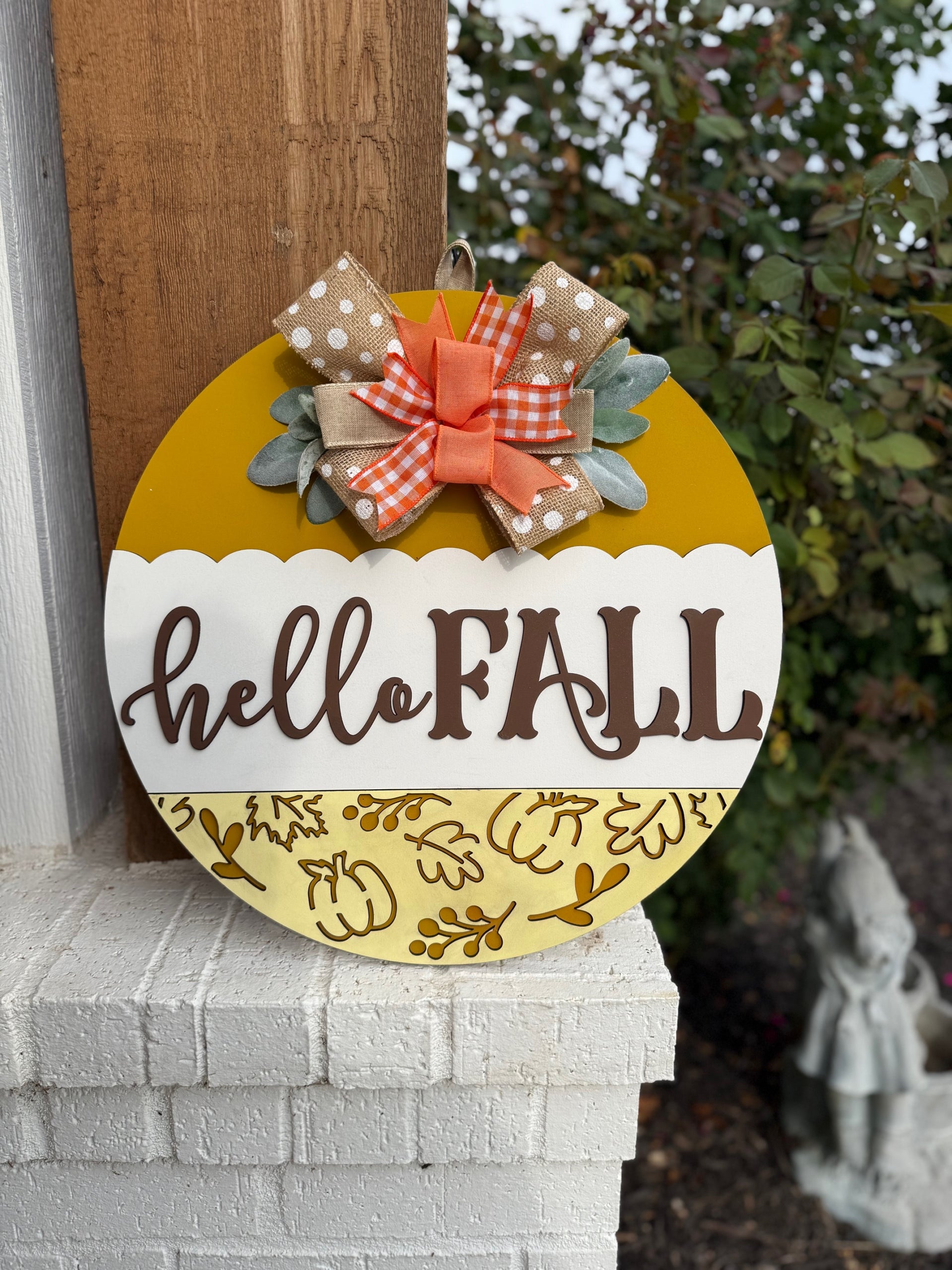 A round yellow and white sign with hello FALL written on it, decorated with a polka dot and plaid bow, autumn leaves, and placed on a white brick ledge next to a wooden post and some greenery.