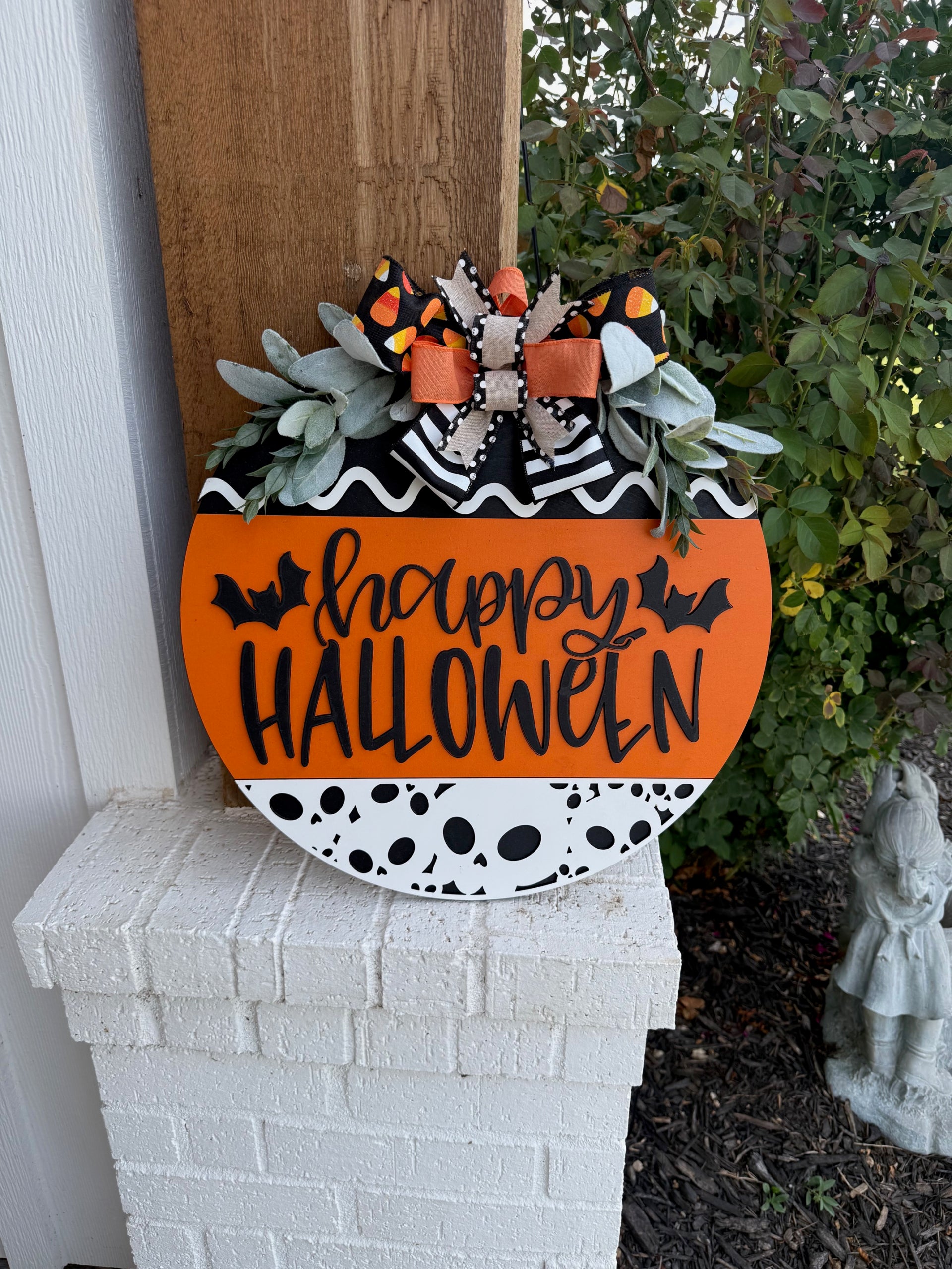 A round Halloween sign with happy HALLOWEEN in black letters, decorated with bats, black-and-white patterns, orange accents, faux greenery, and a large bow with candy corn print at the top.
