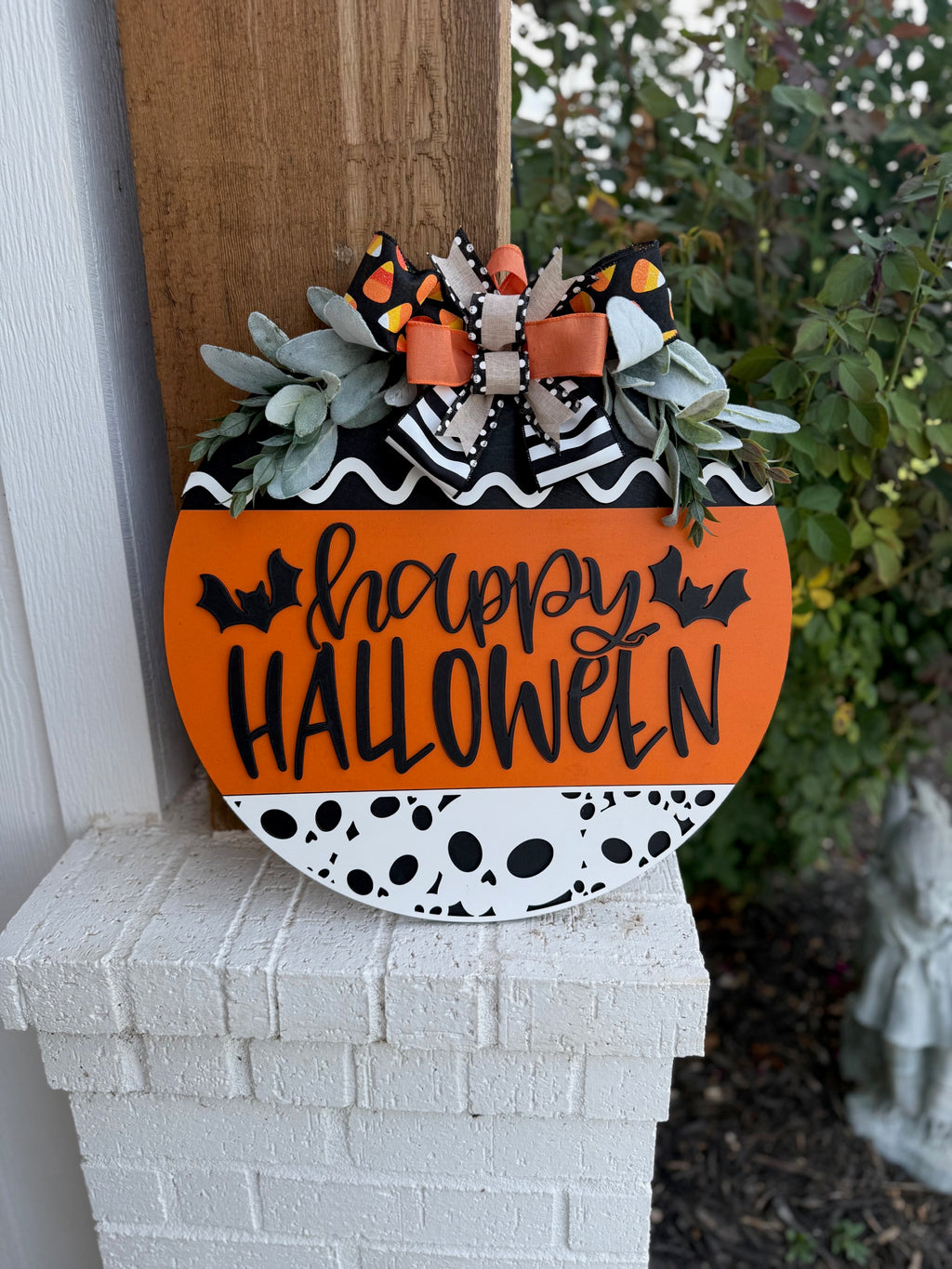 A round Halloween sign with happy HALLOWEEN in black letters, decorated with bats, black-and-white patterns, orange accents, faux greenery, and a large bow with candy corn print at the top.