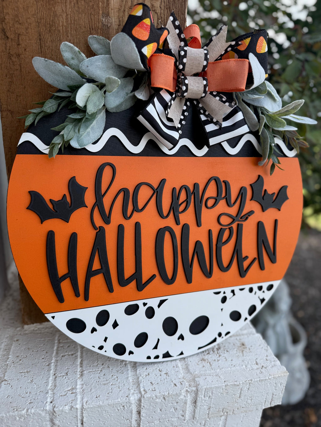 A round Halloween sign with happy HALLOWEEN in black letters, decorated with bats, black-and-white patterns, orange accents, faux greenery, and a large bow with candy corn print at the top.
