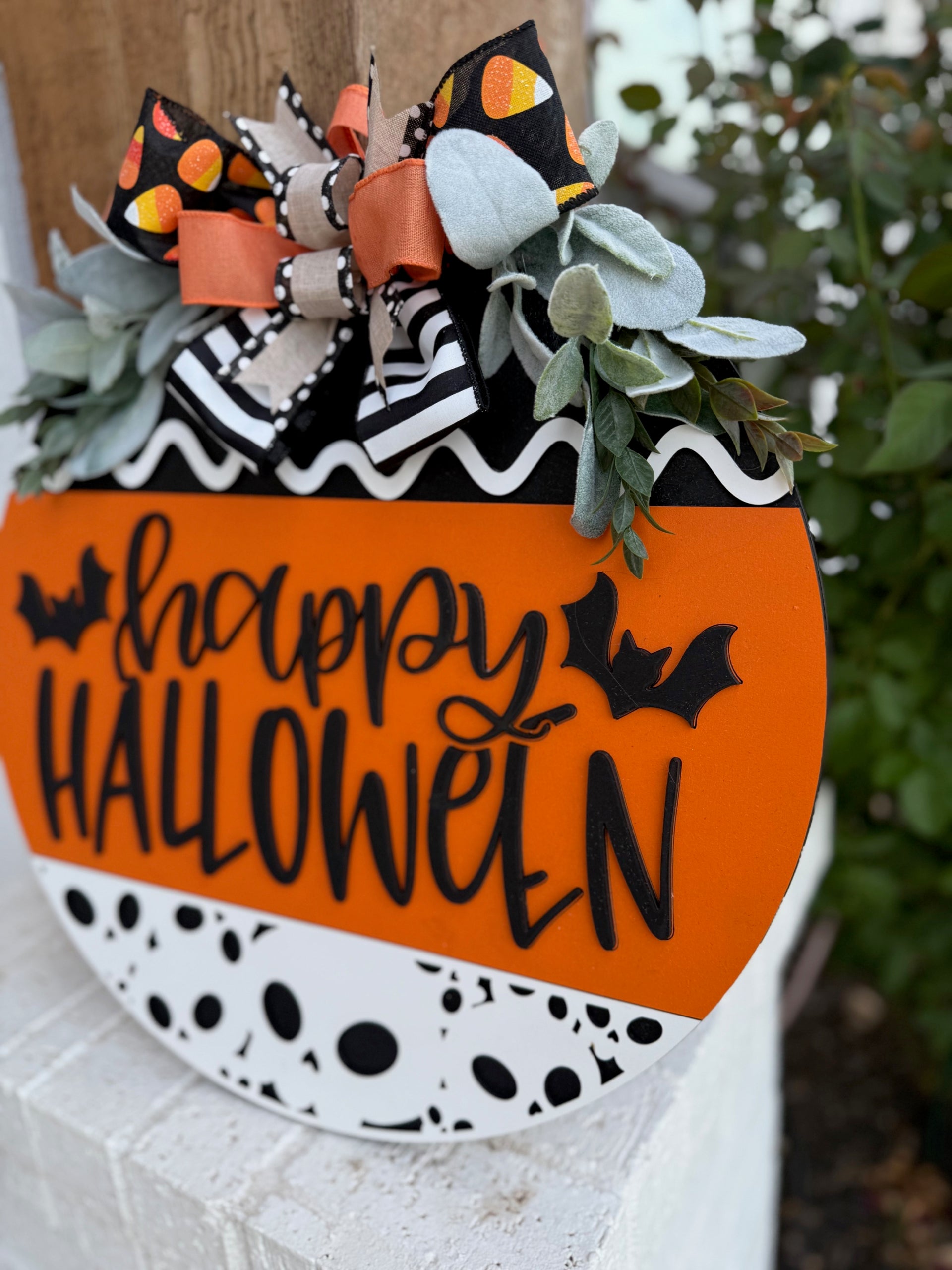 A round Halloween sign with happy HALLOWEEN in black letters, decorated with bats, black-and-white patterns, orange accents, faux greenery, and a large bow with candy corn print at the top.
