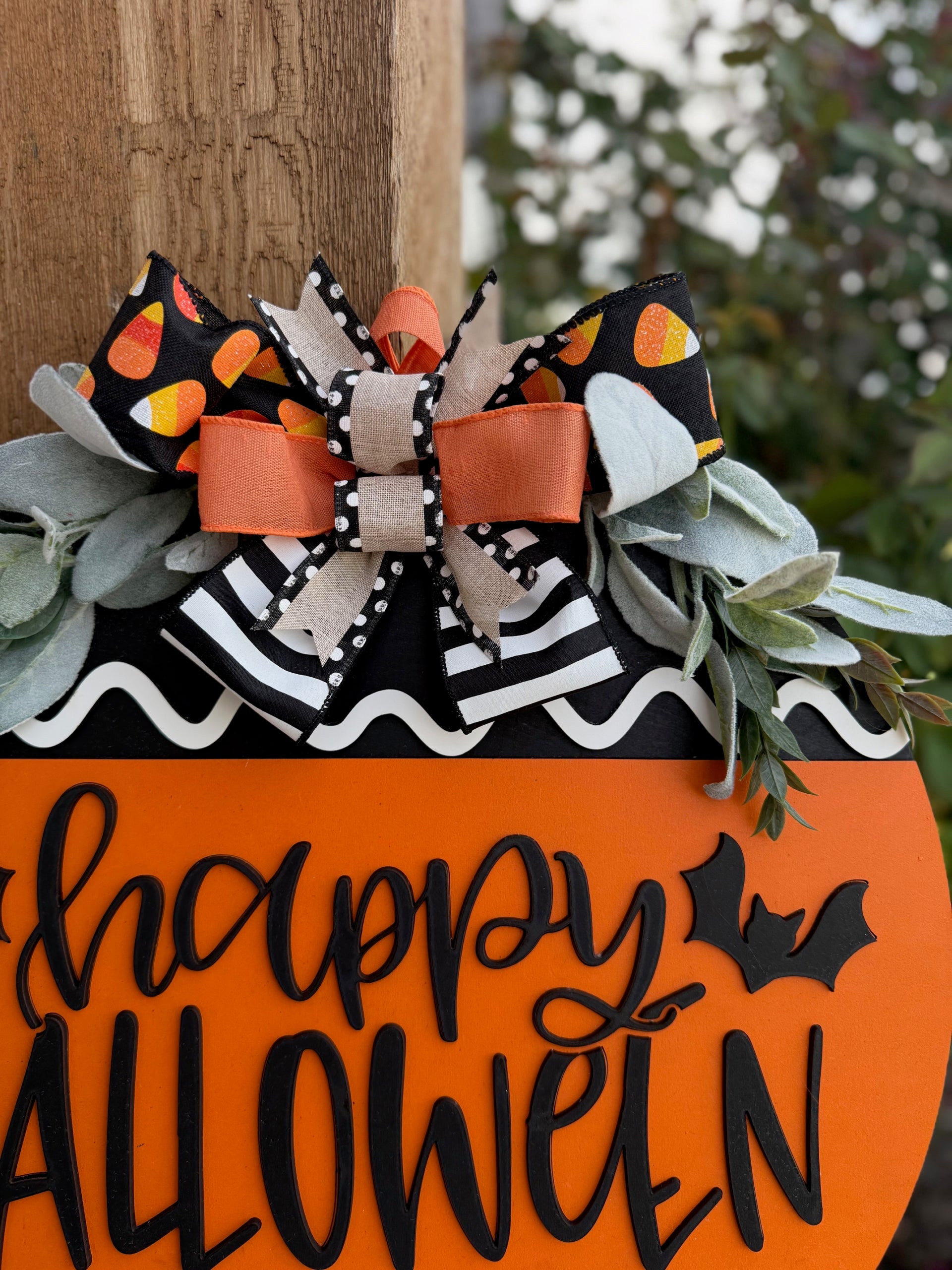 A round Halloween sign with happy HALLOWEEN in black letters, decorated with bats, black-and-white patterns, orange accents, faux greenery, and a large bow with candy corn print at the top.