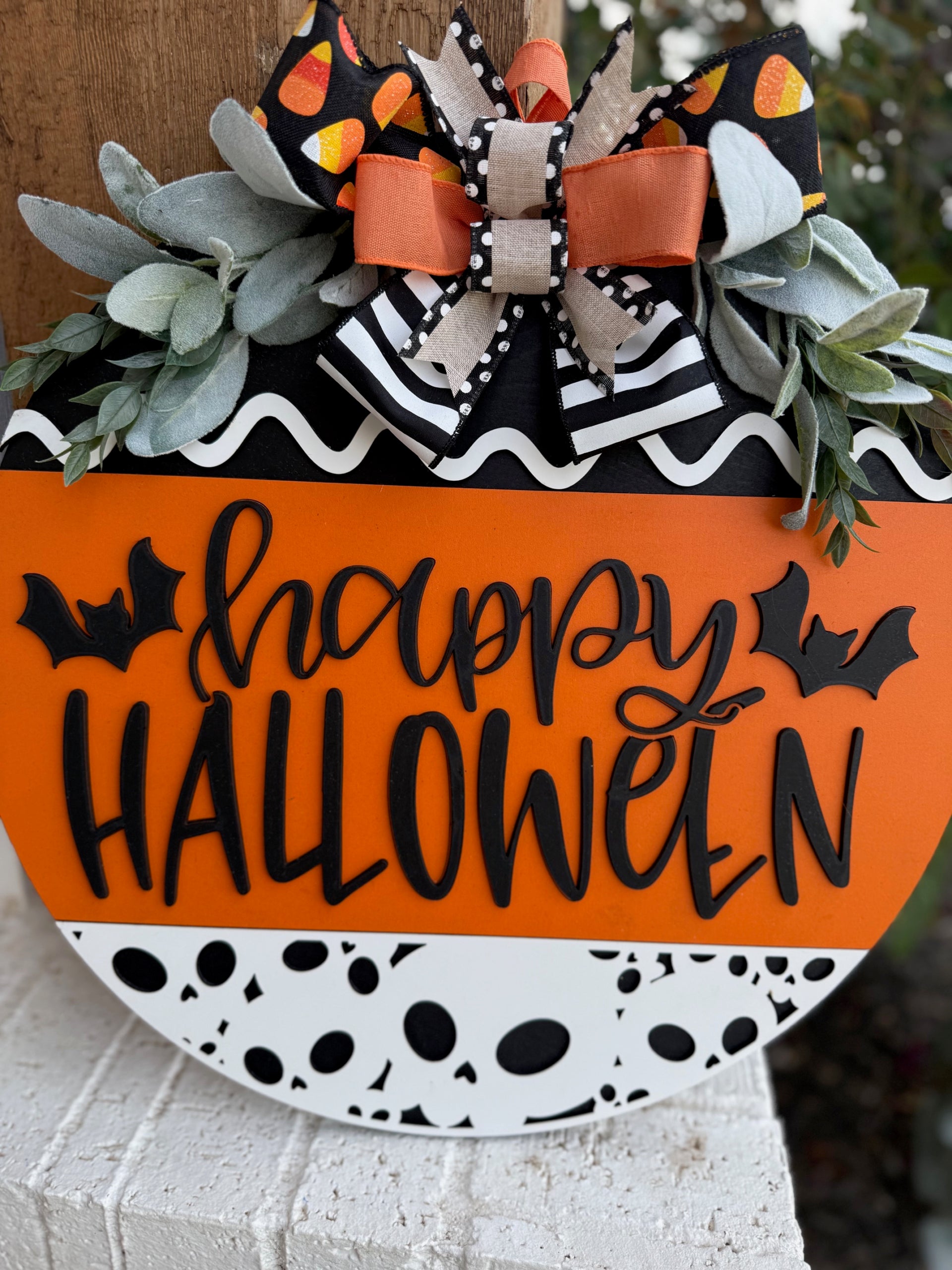 A round Halloween sign with happy HALLOWEEN in black letters, decorated with bats, black-and-white patterns, orange accents, faux greenery, and a large bow with candy corn print at the top.