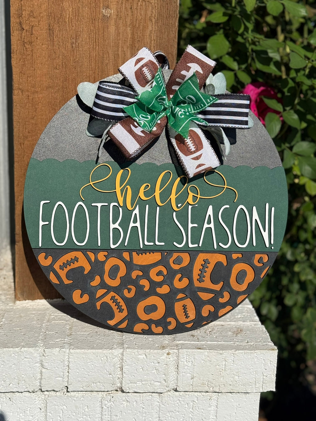 A round wooden sign decorated with a bow at the top reads hello FOOTBALL SEASON! in white and yellow letters, with a pattern of footballs and animal prints on the lower half. The sign is displayed outdoors.