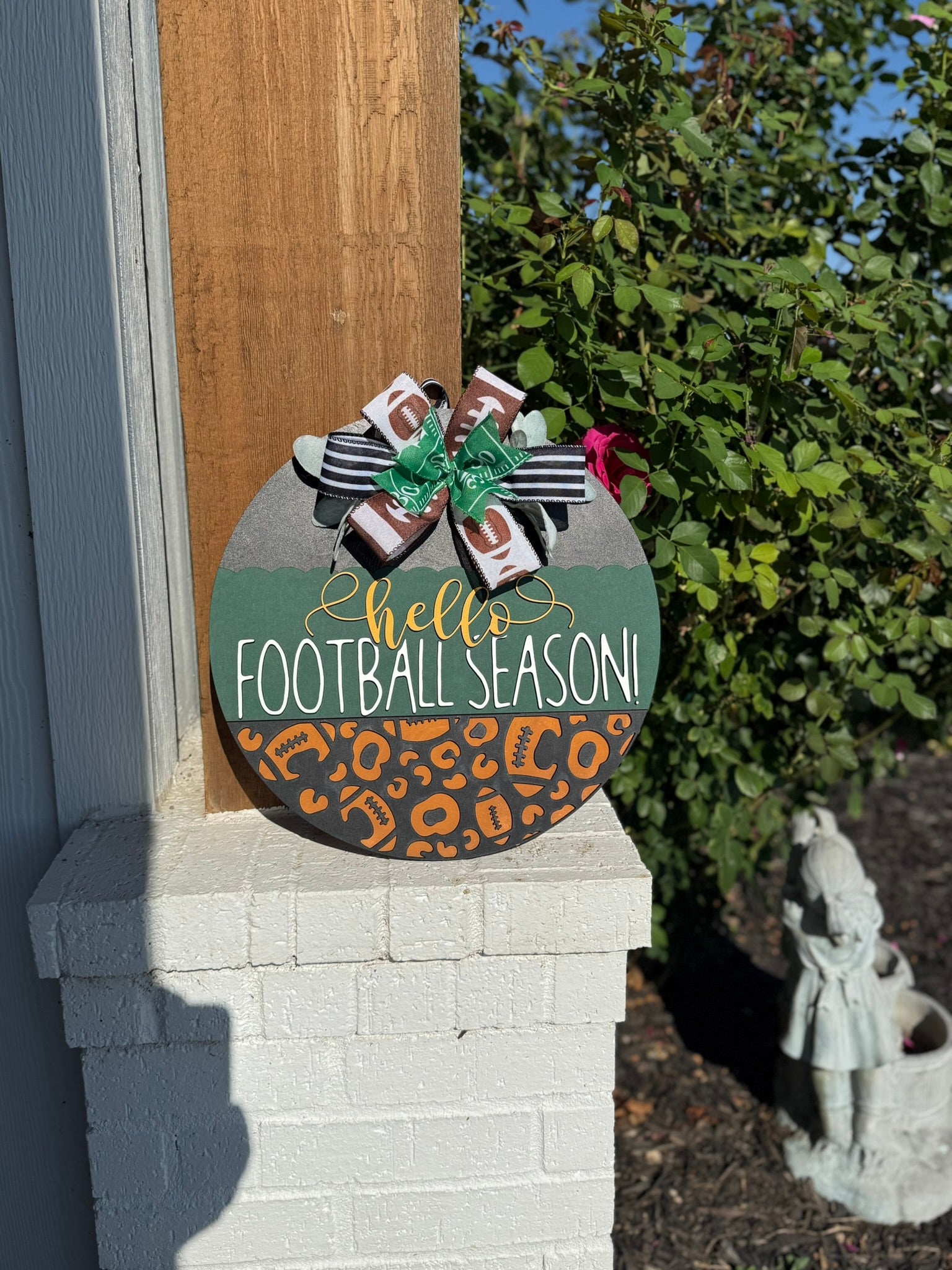 A round wooden sign decorated with a bow at the top reads hello FOOTBALL SEASON! in white and yellow letters, with a pattern of footballs and animal prints on the lower half. The sign is displayed outdoors.