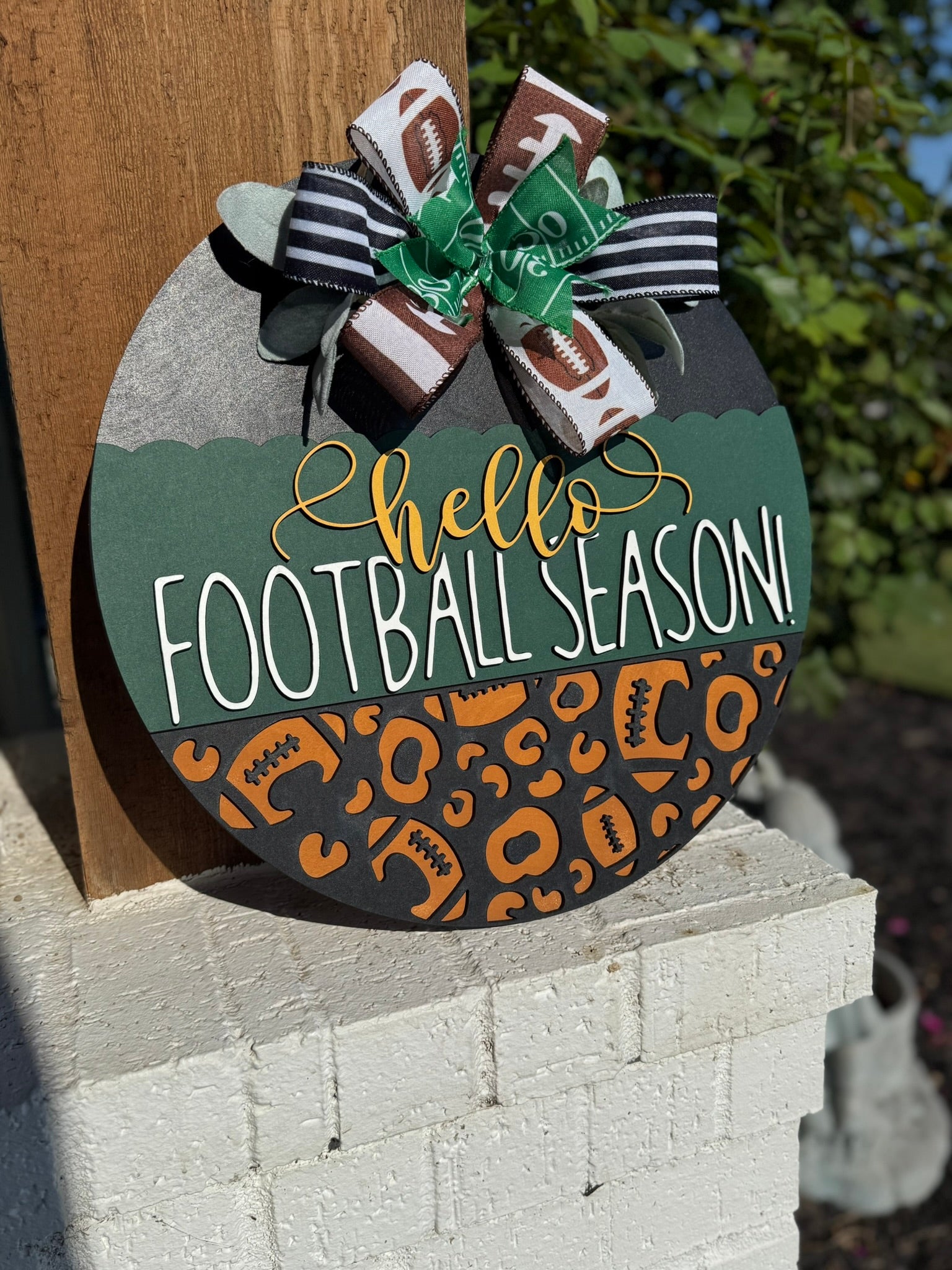 A round wooden sign decorated with a bow at the top reads hello FOOTBALL SEASON! in white and yellow letters, with a pattern of footballs and animal prints on the lower half. The sign is displayed outdoors.