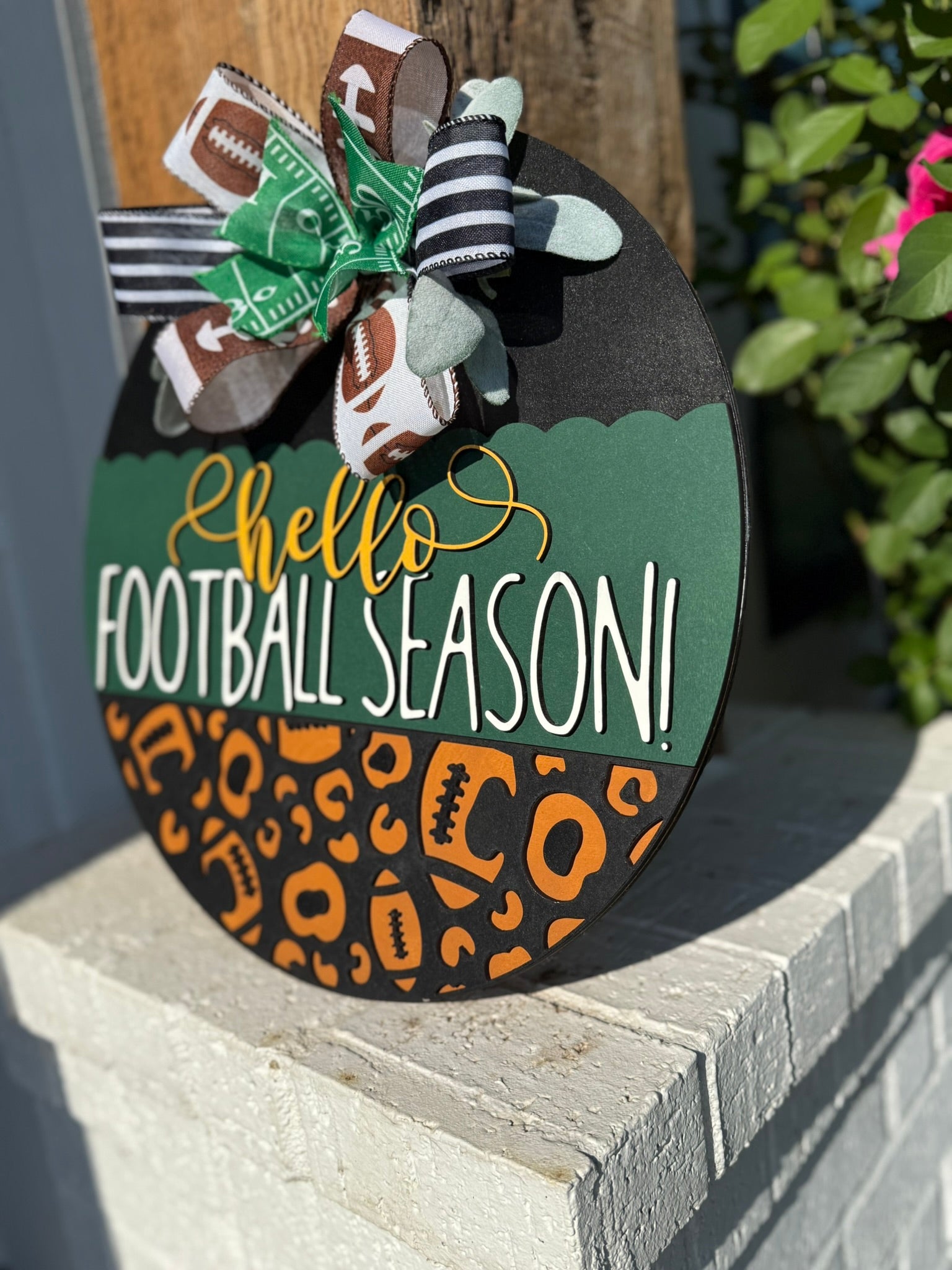 A round wooden sign decorated with a bow at the top reads hello FOOTBALL SEASON! in white and yellow letters, with a pattern of footballs and animal prints on the lower half. The sign is displayed outdoors.