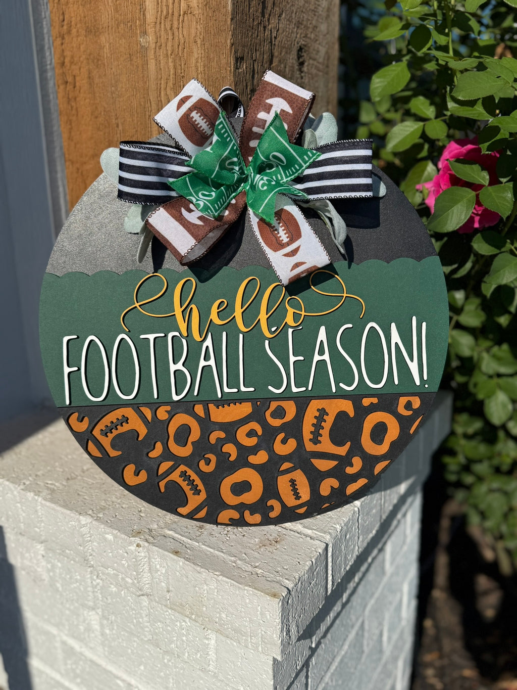 A round wooden sign decorated with a bow at the top reads hello FOOTBALL SEASON! in white and yellow letters, with a pattern of footballs and animal prints on the lower half. The sign is displayed outdoors.