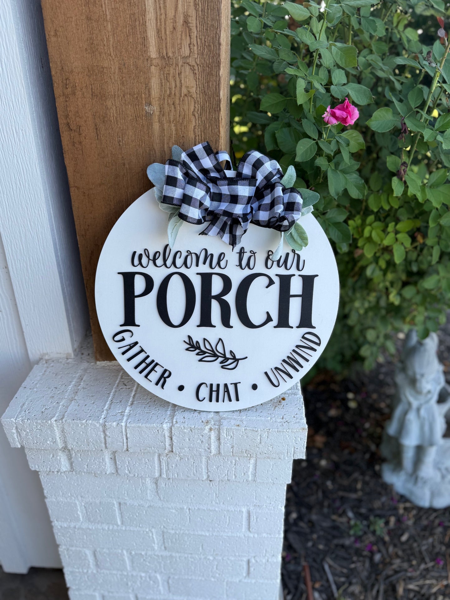 A round sign with a black and white plaid bow reads Welcome to our porch - Gather • Chat • Unwind. It leans against a wooden post and white brick, with a rose bush and garden statue in the background.