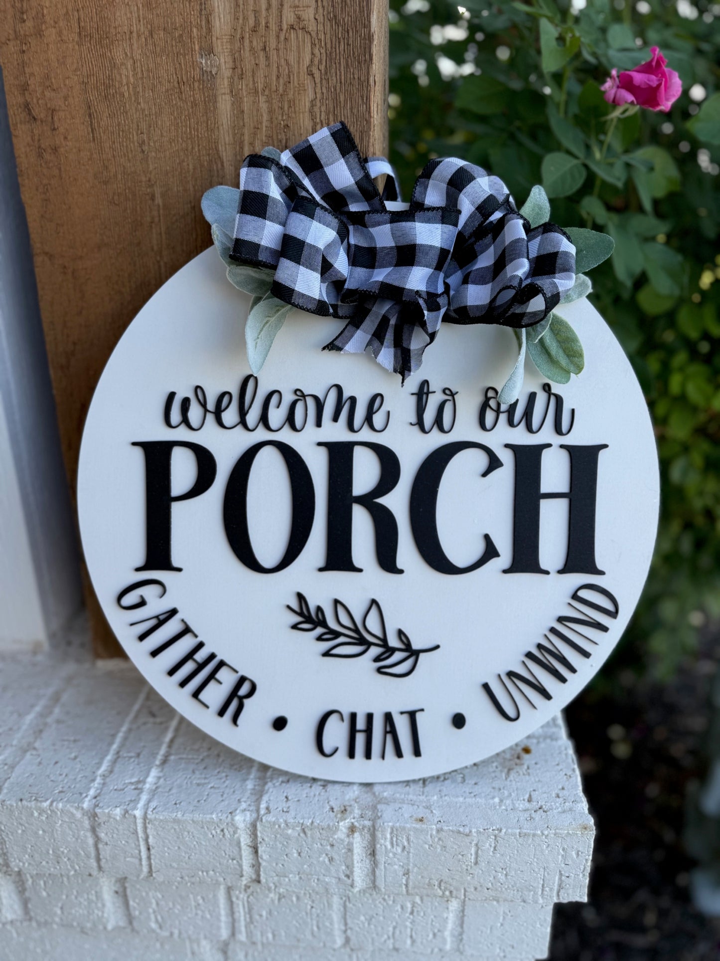 A round white porch sign with black text reads, “Welcome to our porch. Gather, chat, unwind.” It is decorated with a black-and-white plaid bow and green leaves, leaning against a brick wall near flowers.