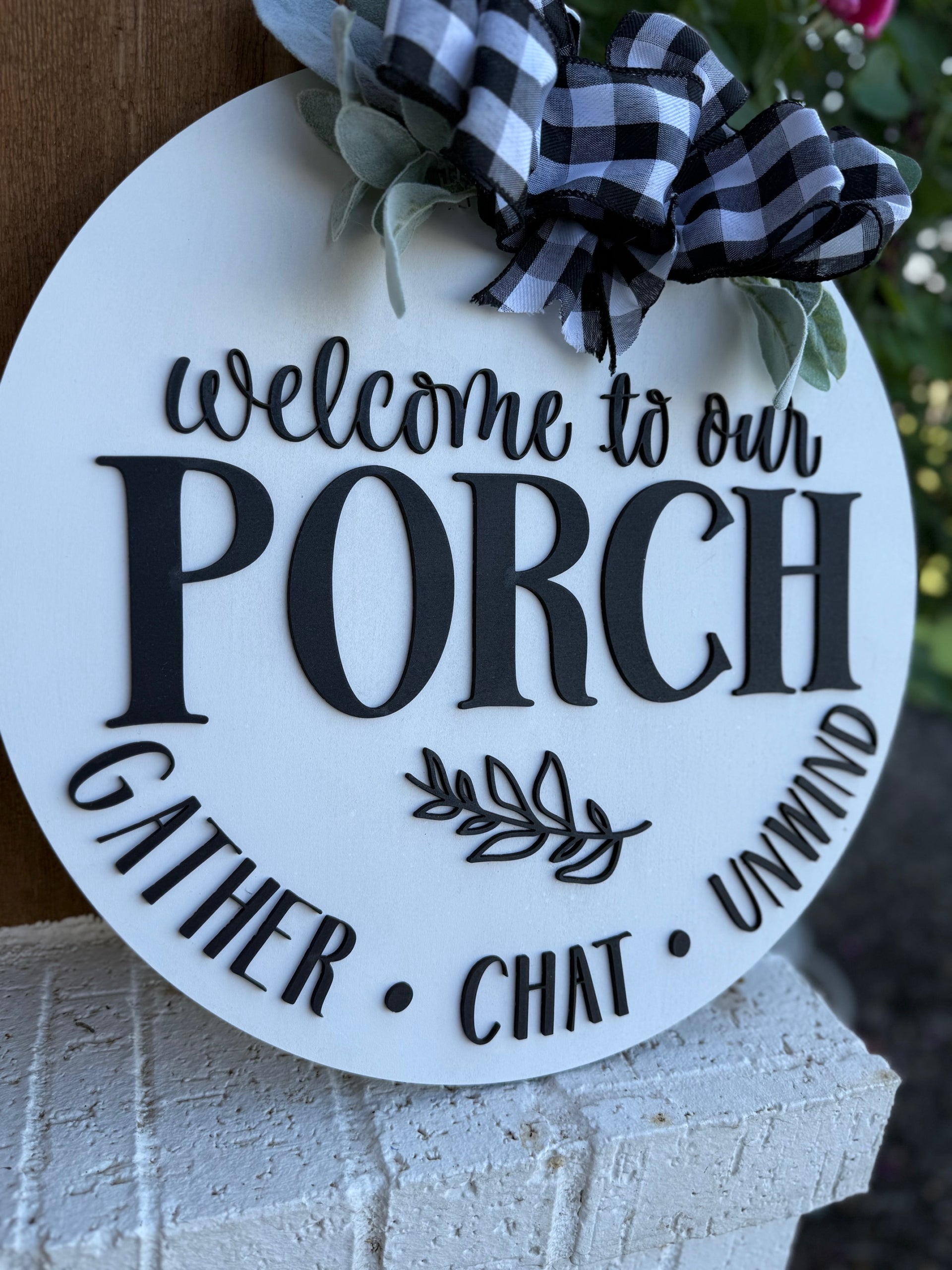 A round white porch sign decorated with a black and white checkered bow and greenery, featuring the text: Welcome to our porch. Gather • Chat • Unwind.