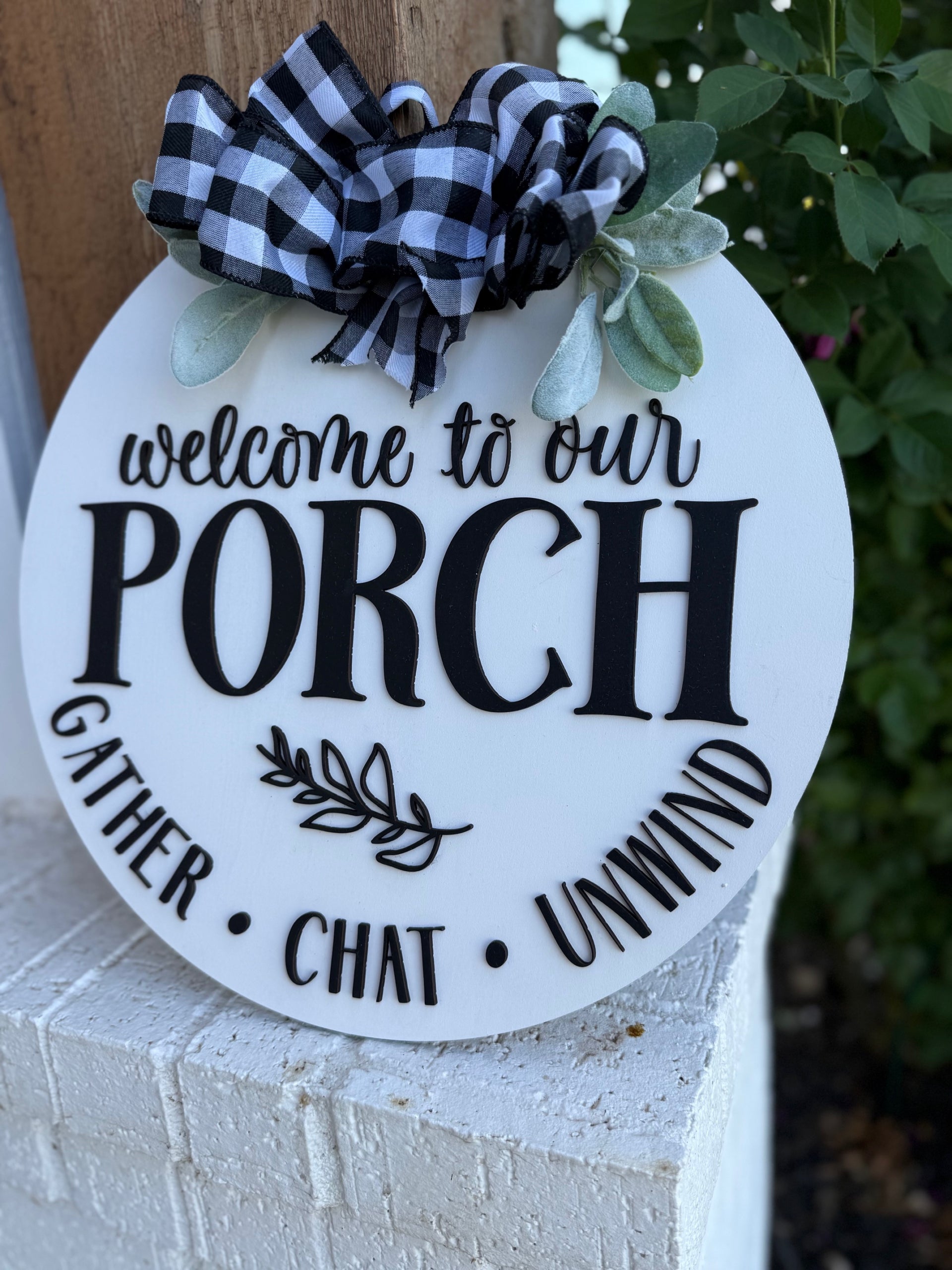 A round welcome sign on a white brick surface reads “Welcome to our Porch” with Gather, Chat, and Unwind around the edge. It is decorated with black-and-white plaid ribbon and greenery.