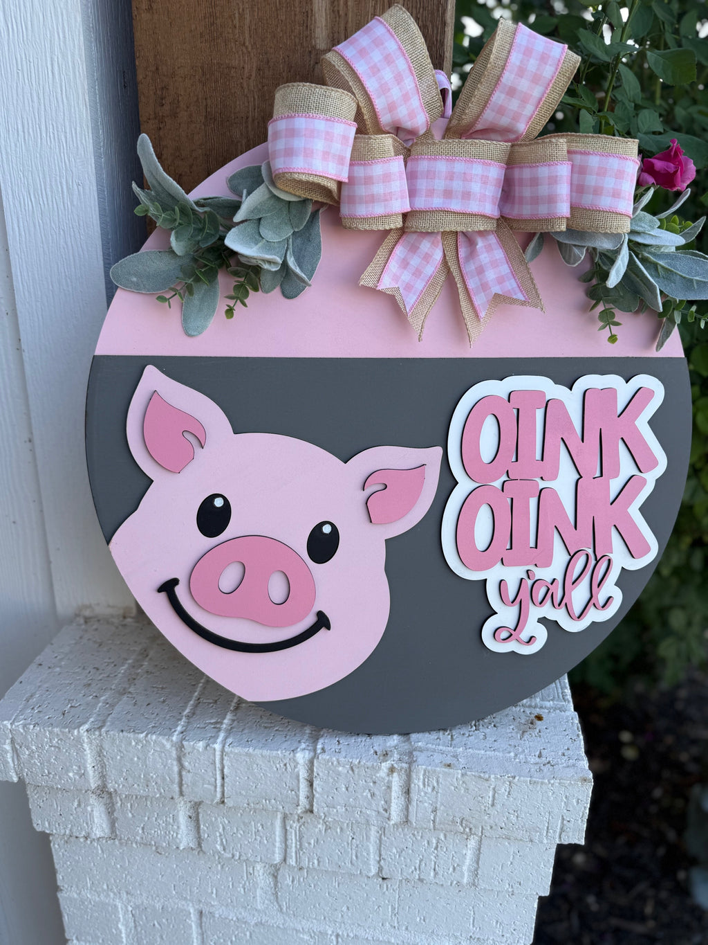 A round decorative sign with a smiling pink pig, pink and white bow, green leaves, and the words OINK OINK yall in white letters, displayed on a white brick surface.
