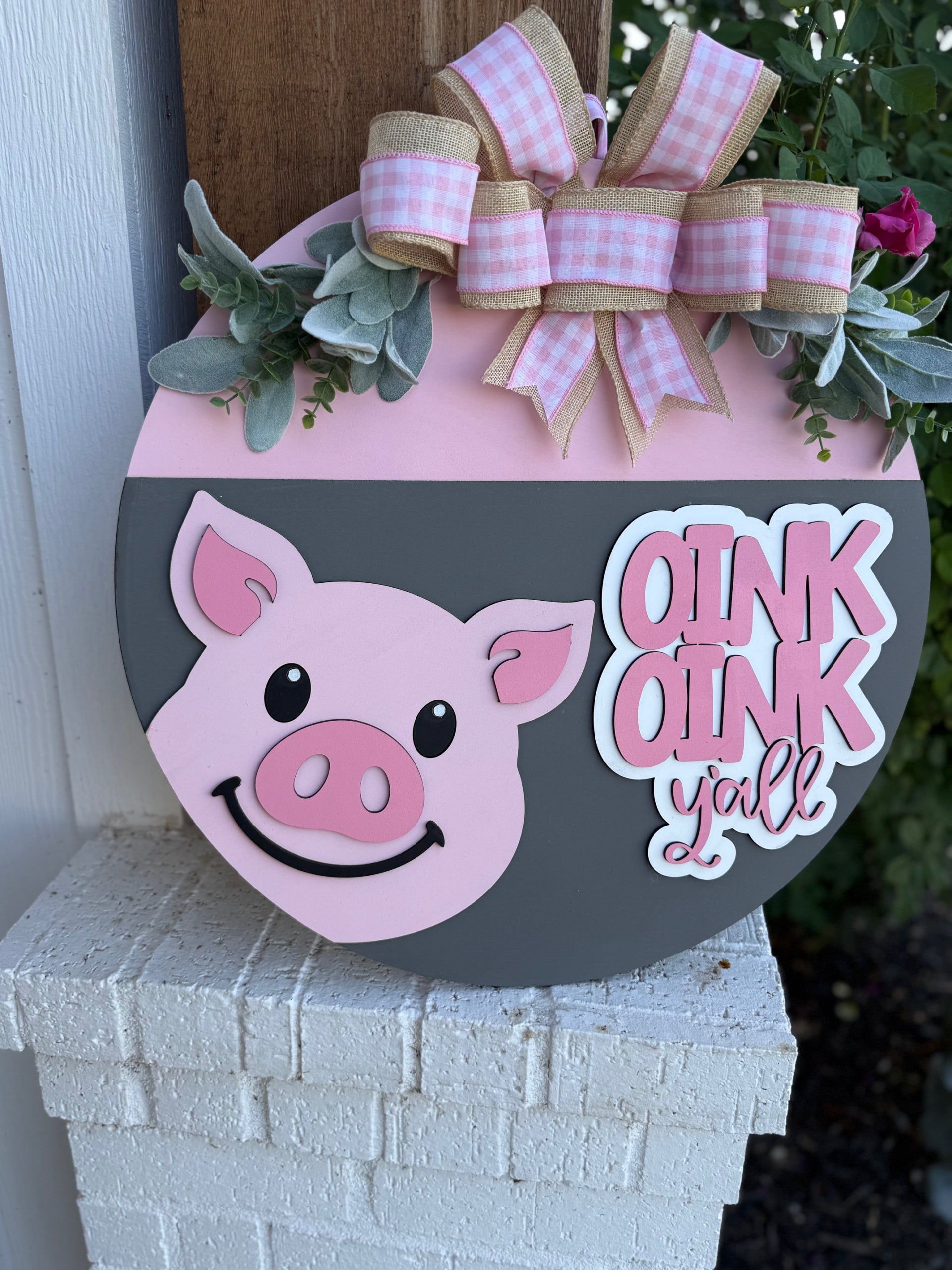 A round decorative sign with a smiling pink pig, pink and white bow, green leaves, and the words OINK OINK yall in white letters, displayed on a white brick surface.