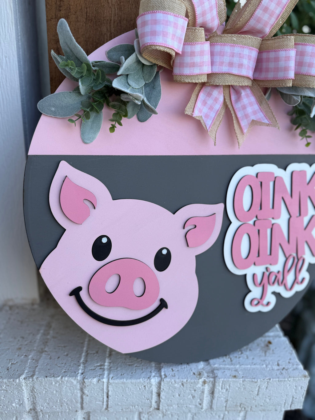 A round welcome sign with a smiling pink pig face, green leaves, and a pink gingham bow. Text on the sign reads “OINK OINK y’all.” The sign is displayed outdoors on a brick ledge.