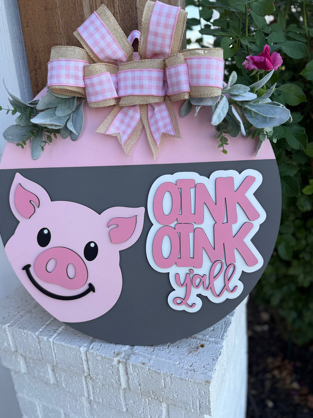 A round decorative sign featuring a smiling pink pig face, pink and white plaid bows, greenery accents, and the words OINK OINK yall in playful lettering. The sign rests on a white brick surface by flowers.