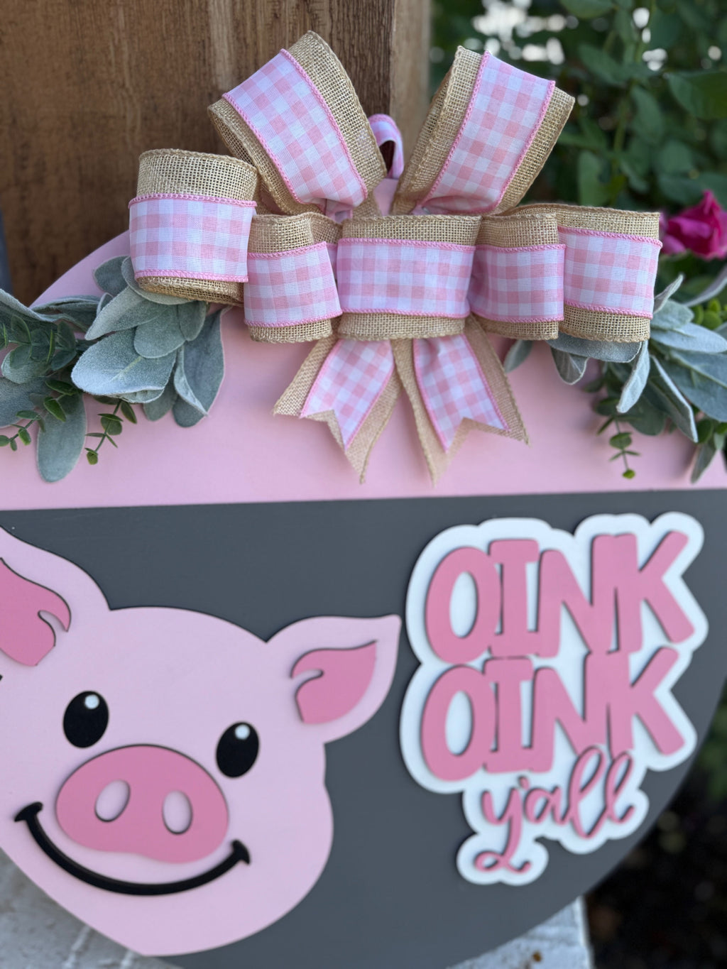 A decorative sign featuring a cute cartoon pig face, pink and burlap plaid bow, green leaves, and the phrase Oink Oink y’all in playful white and pink lettering.