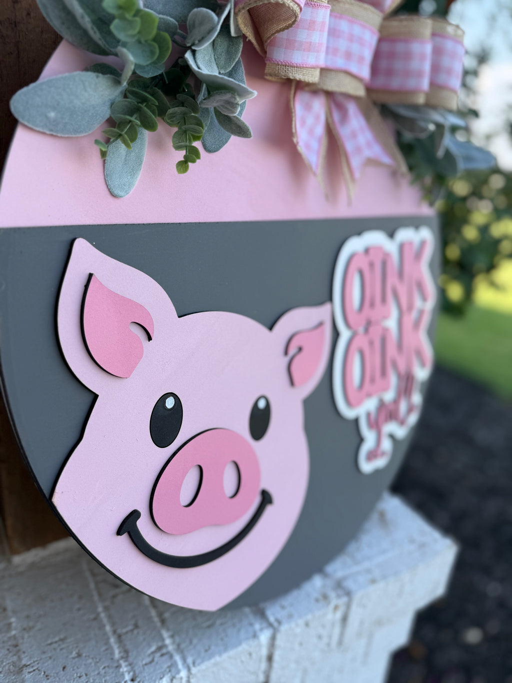 A decorative sign features a smiling pink pig face and the words “OINK OINK” with greenery and a pink plaid bow on top, hanging outdoors with blurred trees and lawn in the background.