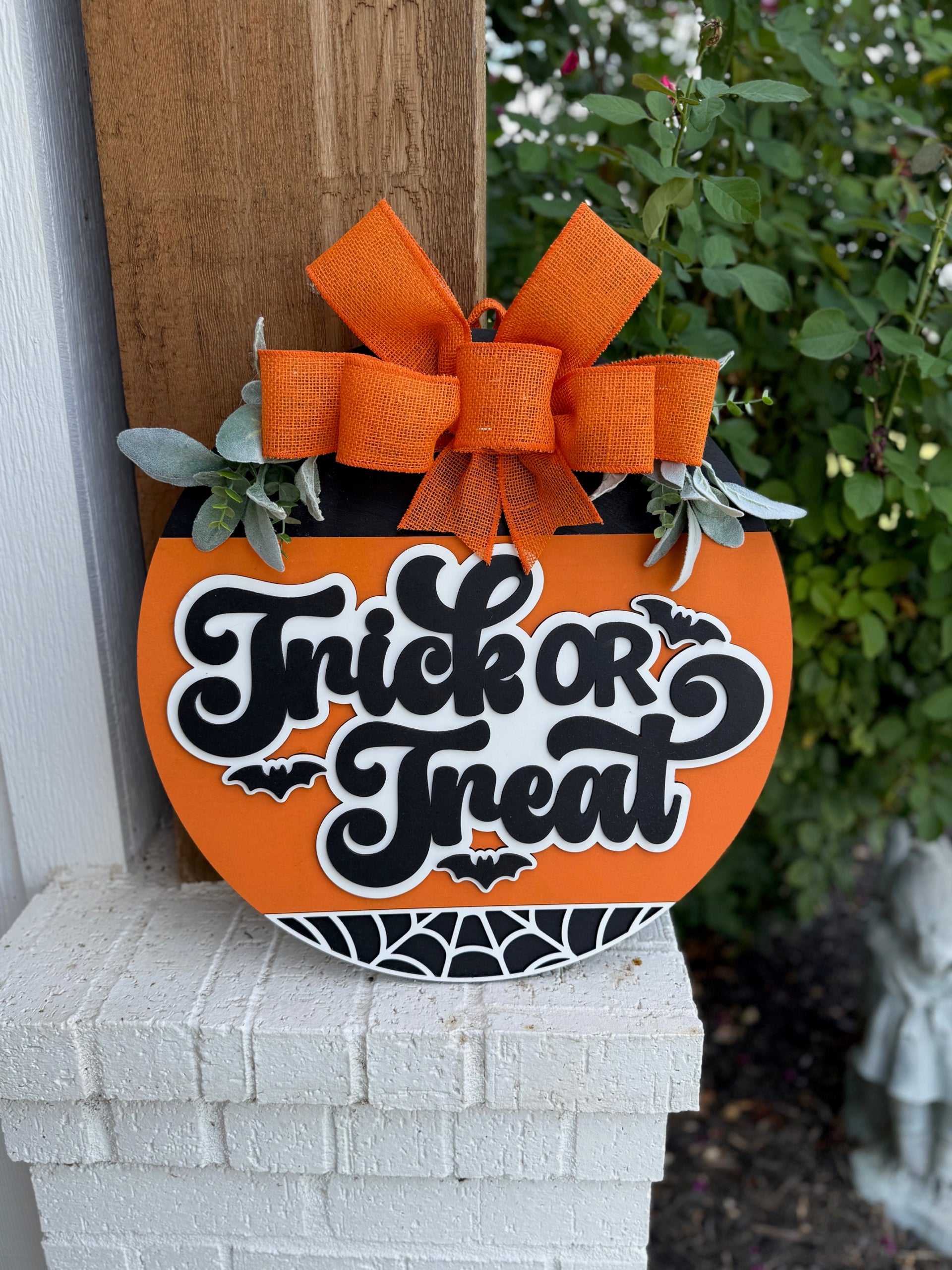 A round orange sign with a large orange bow and green leaves reads Trick or Treat in black and white letters, decorated with bat and spider web designs, hanging on a wooden post near white brick and greenery.