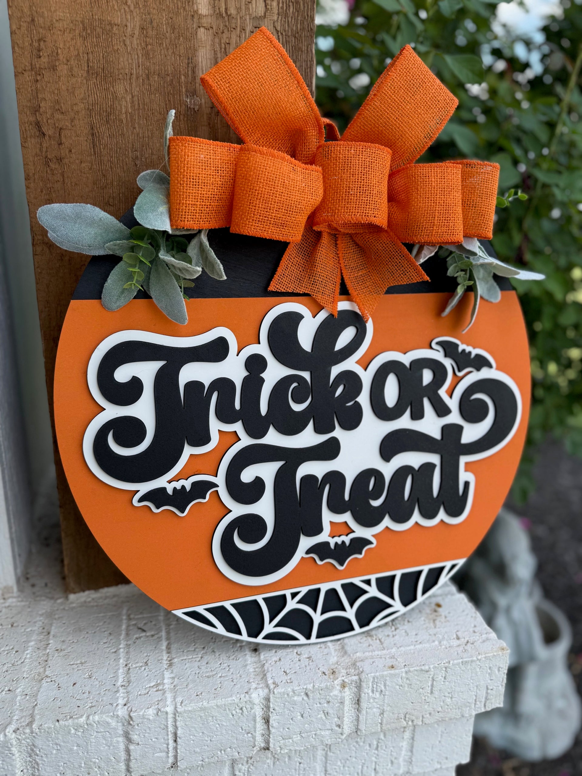 A round orange sign shaped like a pumpkin, decorated with a large orange bow, green leaves, and the words Trick or Treat in bold letters, surrounded by small bats and a spiderweb design at the bottom.