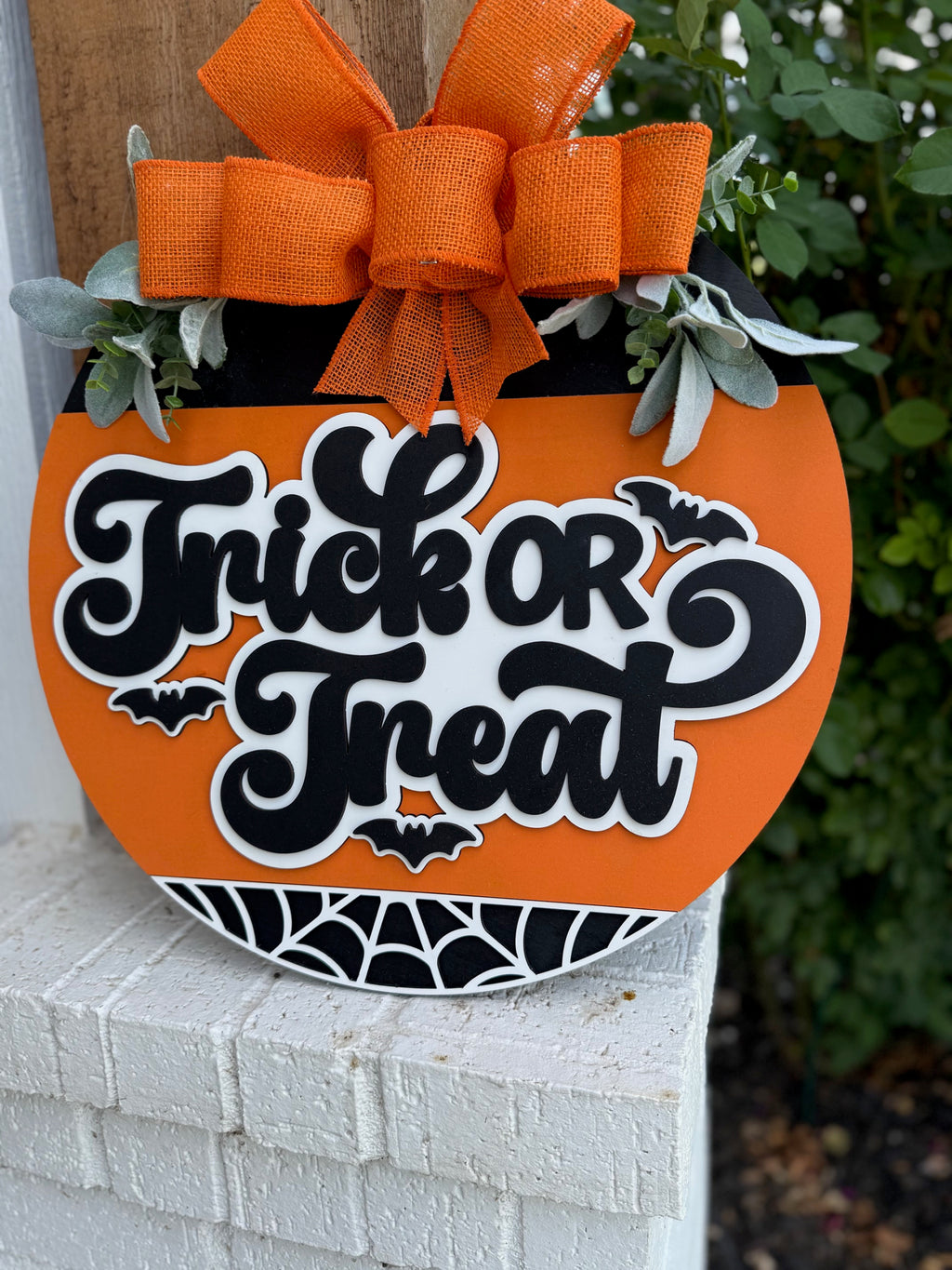 A round orange Halloween sign with the words Trick or Treat in bold black and white letters, decorated with bats, a spiderweb design, faux greenery, and topped with a large orange ribbon bow.