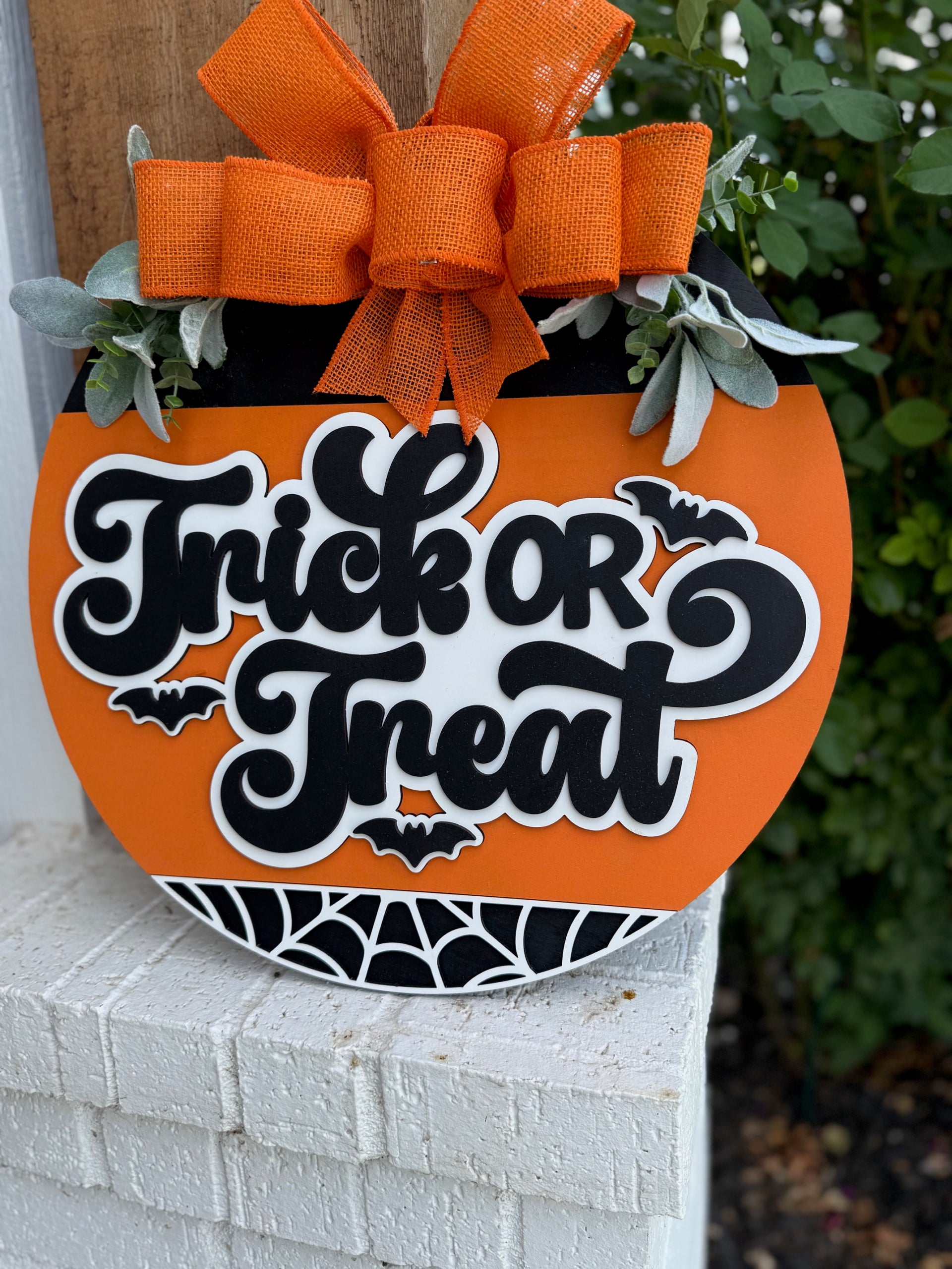 A round orange Halloween sign with the words Trick or Treat in bold black and white letters, decorated with bats, a spiderweb design, faux greenery, and topped with a large orange ribbon bow.