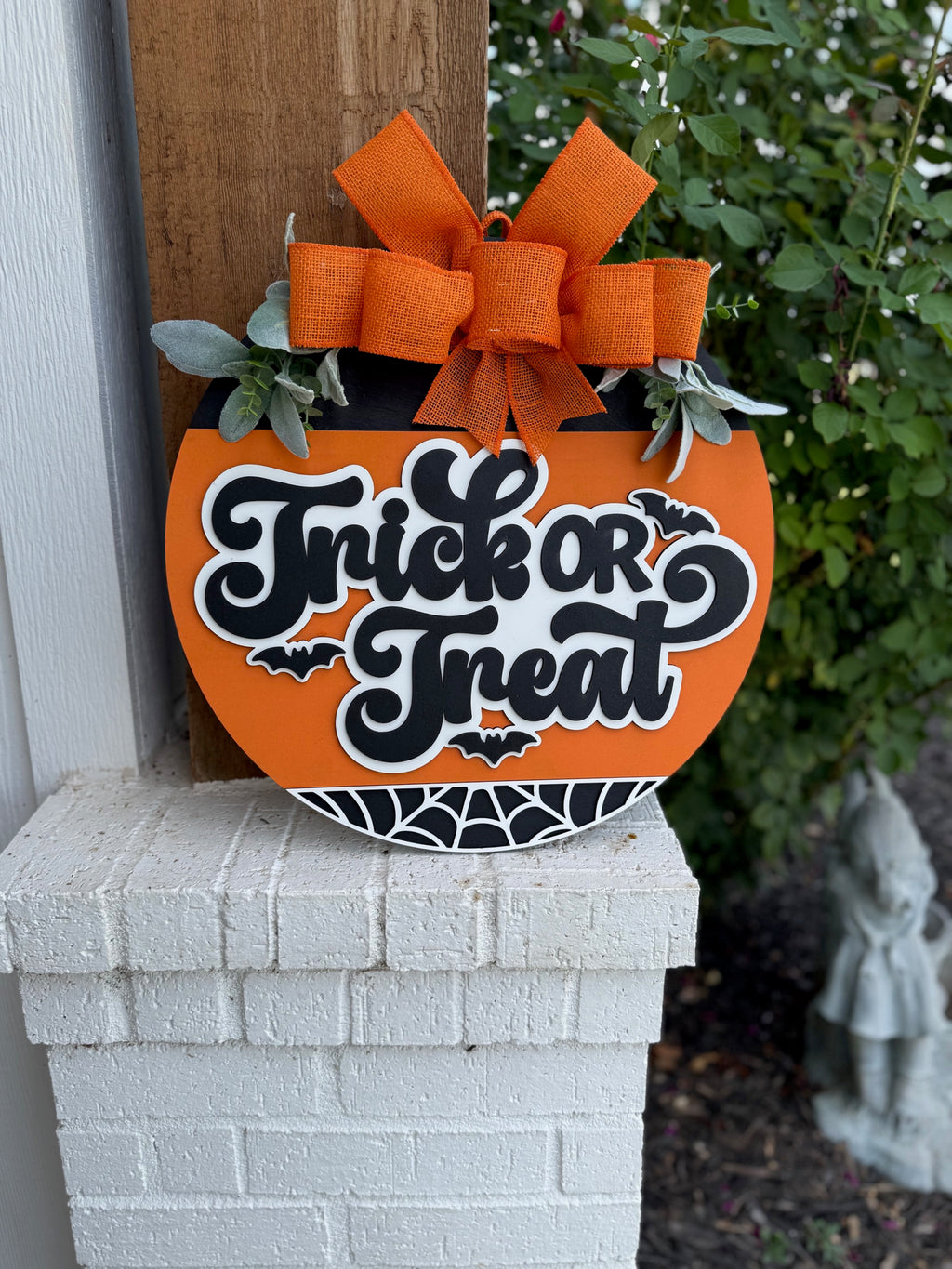 An orange pumpkin-shaped sign with a large orange bow and greenery reads Trick or Treat in bold black and white letters, decorated with bats and a spiderweb, resting on a white brick ledge near green plants.