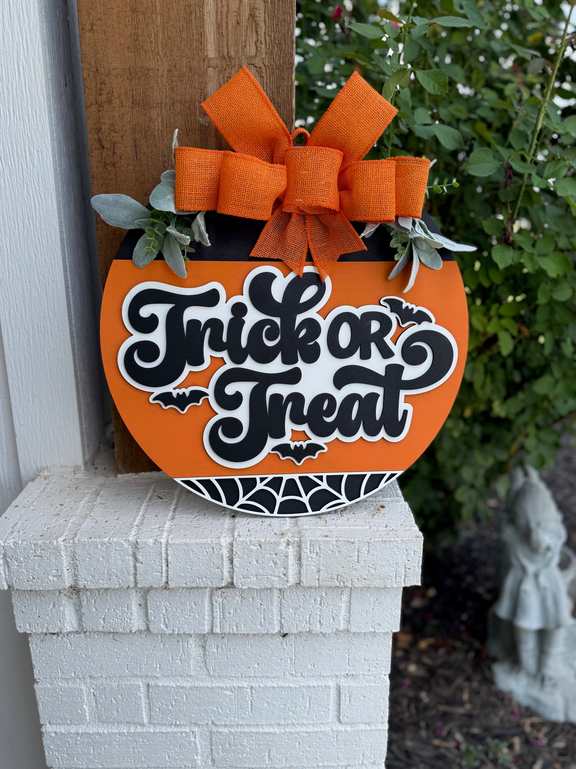 An orange pumpkin-shaped sign with a large orange bow and greenery reads Trick or Treat in bold black and white letters, decorated with bats and a spiderweb, resting on a white brick ledge near green plants.