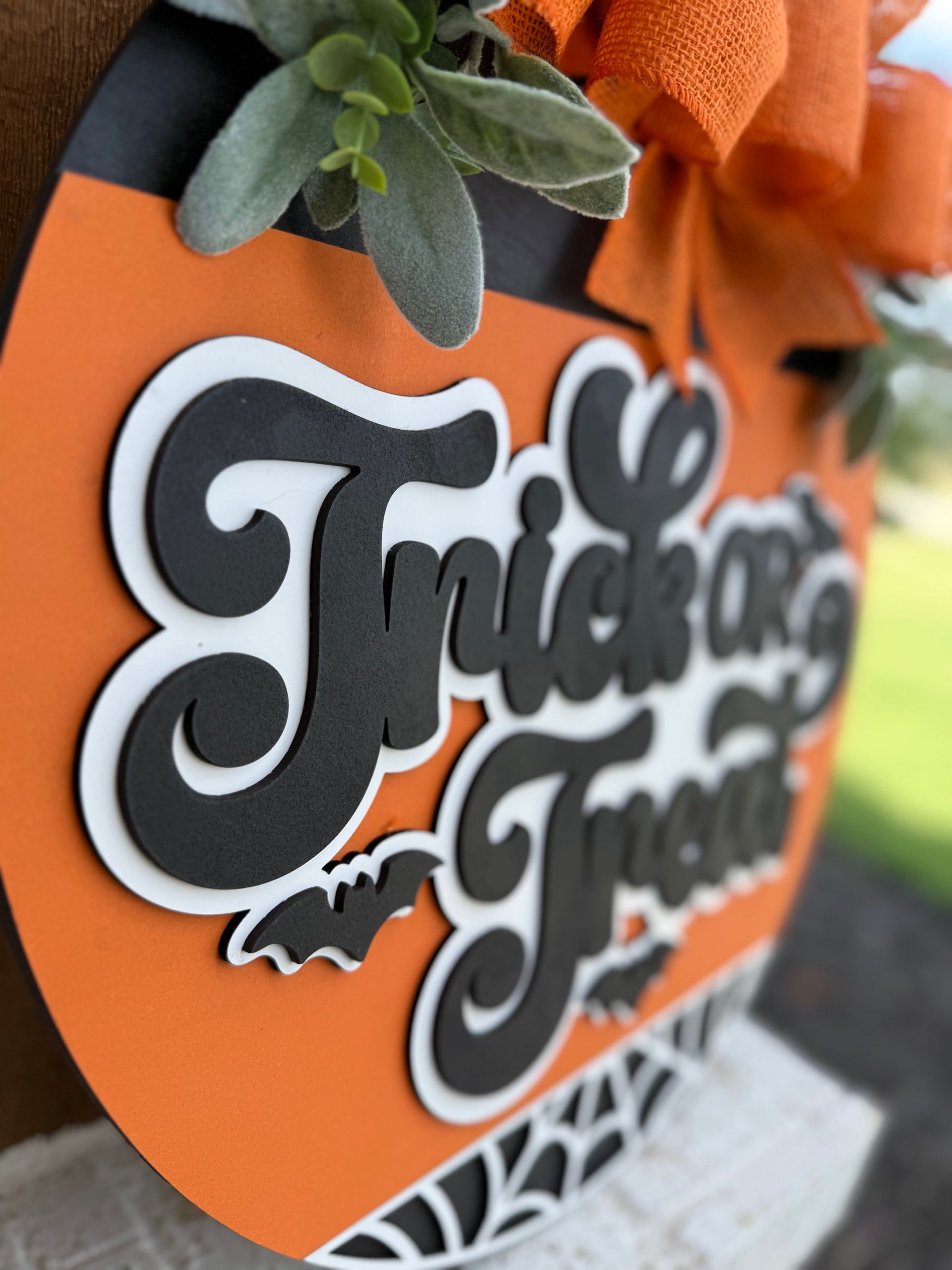 Close-up of a round, orange Halloween sign with black and white layered text reading Trick or Treat, decorated with green leaves and an orange bow. A small bat shape is below the text.