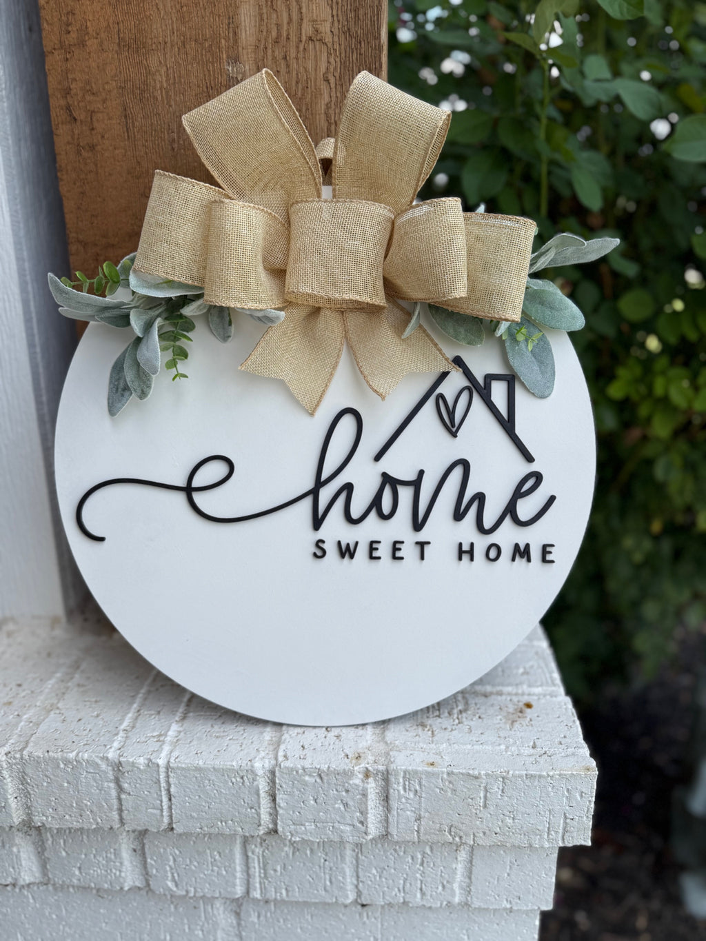 A round white sign with “home sweet home” in black script, decorated with a burlap bow and green leaves, hangs on a white brick wall near some green plants.