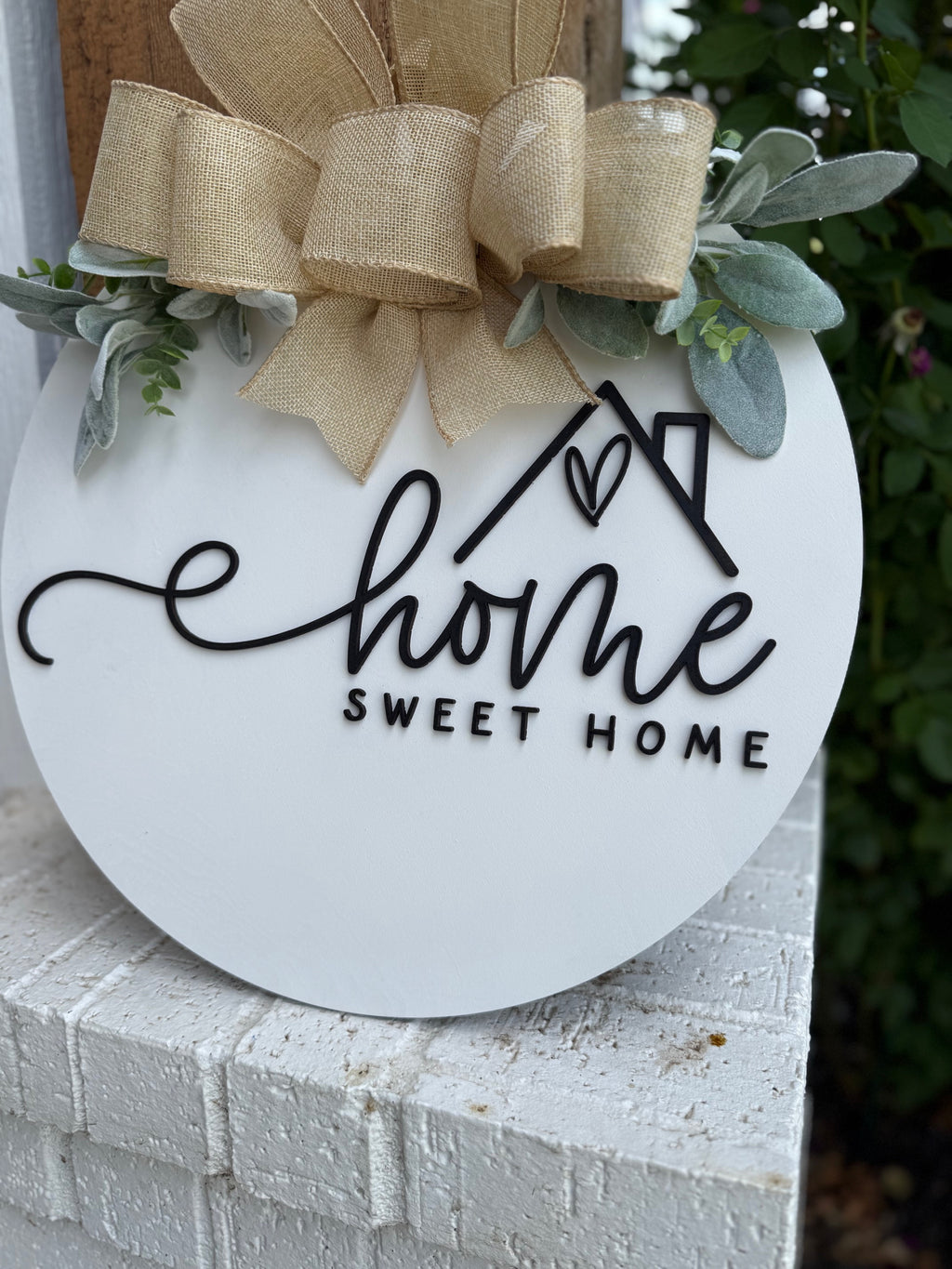 A round white sign with the words home sweet home in black script, decorated with a tan burlap bow and green leaves, sits on a white brick ledge.