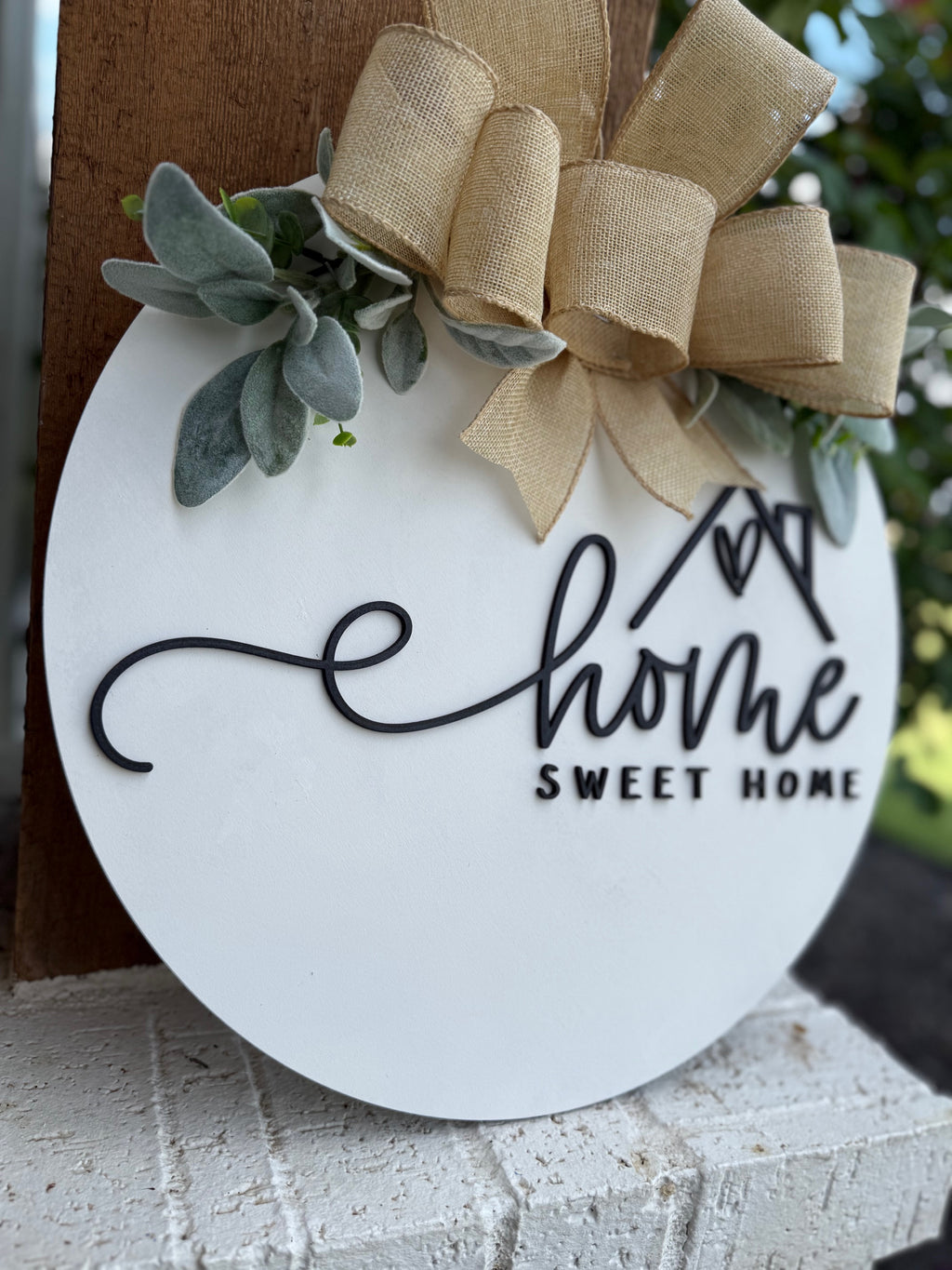 A round white sign with “Home Sweet Home” in black script, decorated with a burlap bow and green leaves, hangs on a wall near a garden.