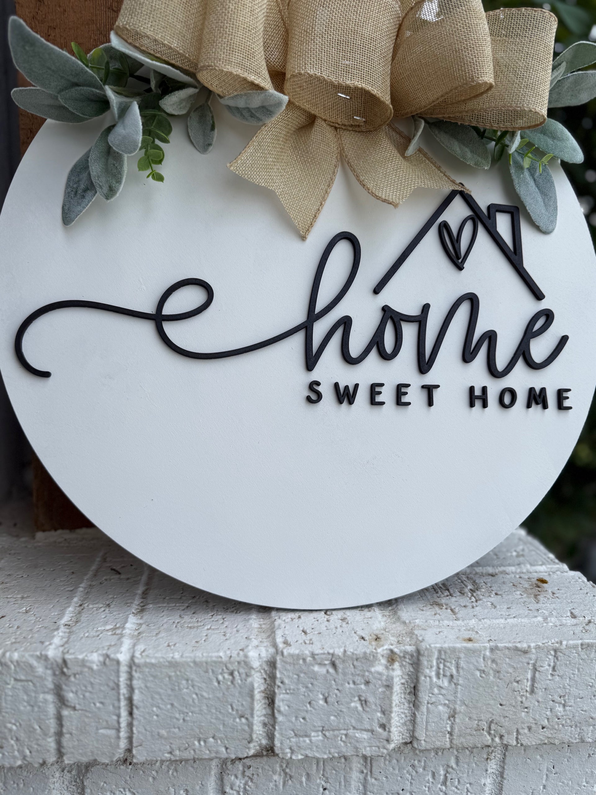 A round white sign with black lettering that says Home Sweet Home, decorated with a tan burlap bow and green foliage at the top, displayed on a white brick surface.