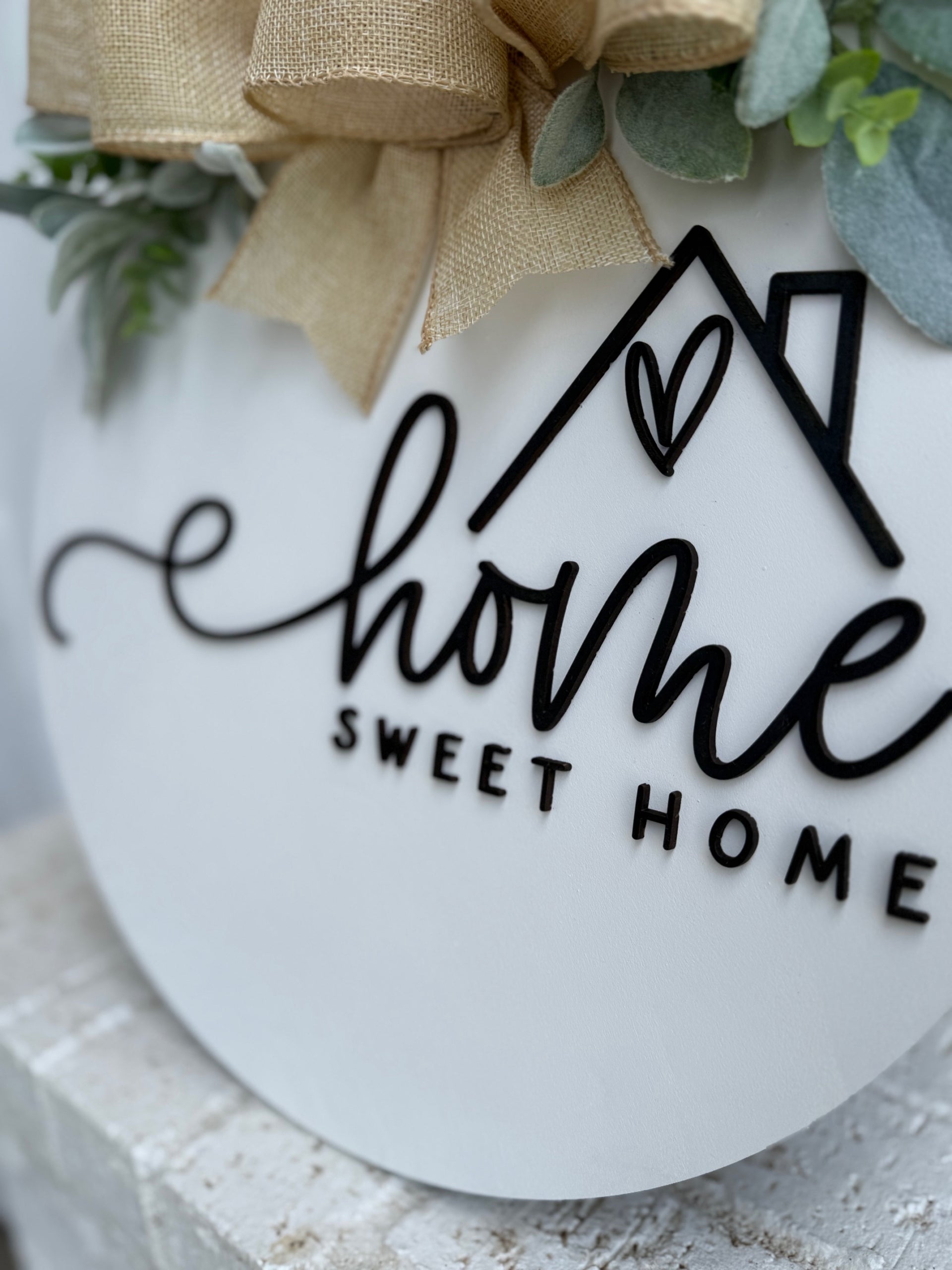 A round white sign decorated with greenery and a burlap bow, featuring the words home sweet home in black lettering with a house outline and heart above the word home.