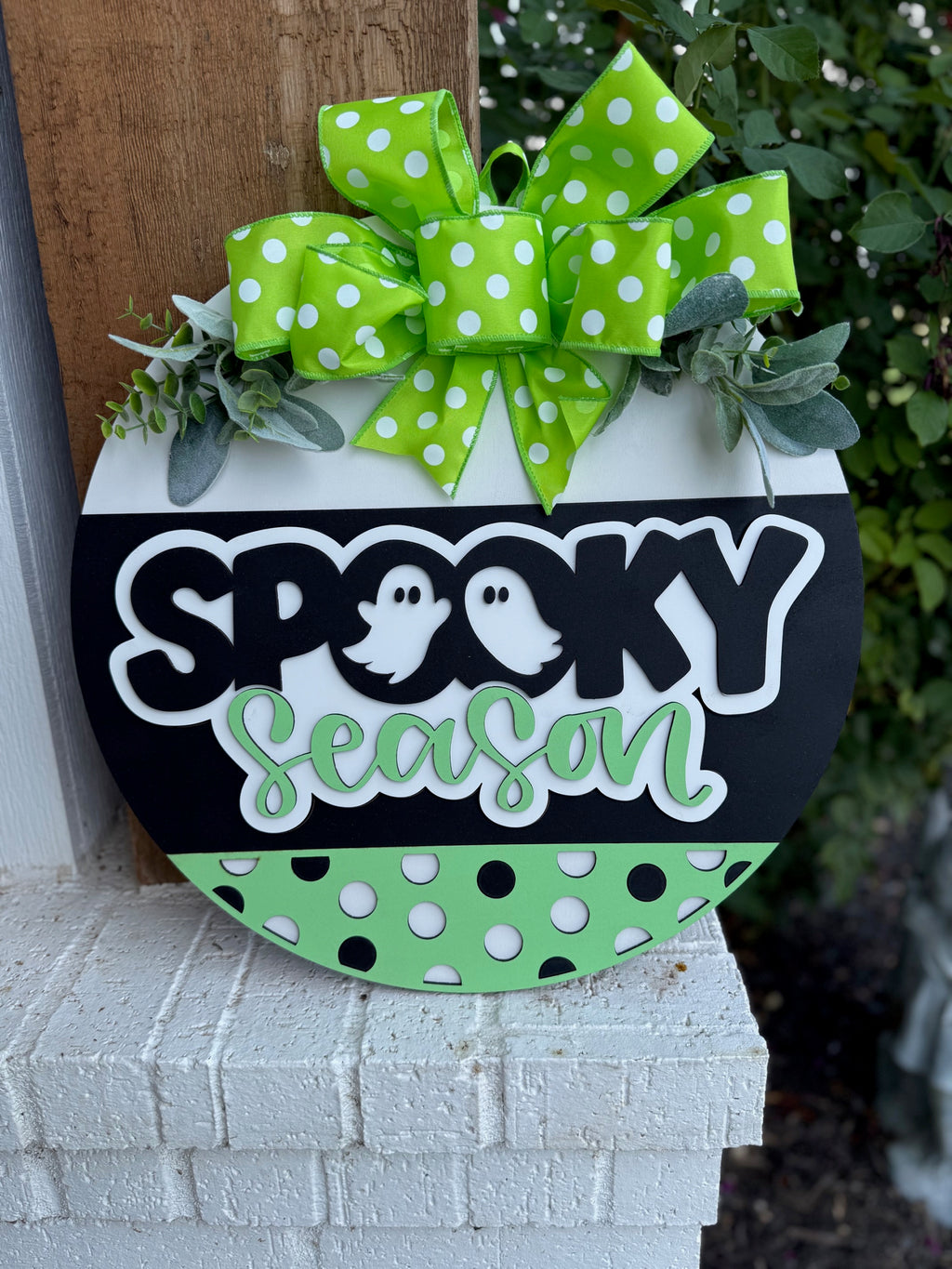 A round sign decorated with a green polka dot bow and greenery, featuring the words “Spooky Season” with ghost illustrations, in black, white, and green colors with polka dot accents, displayed outdoors.