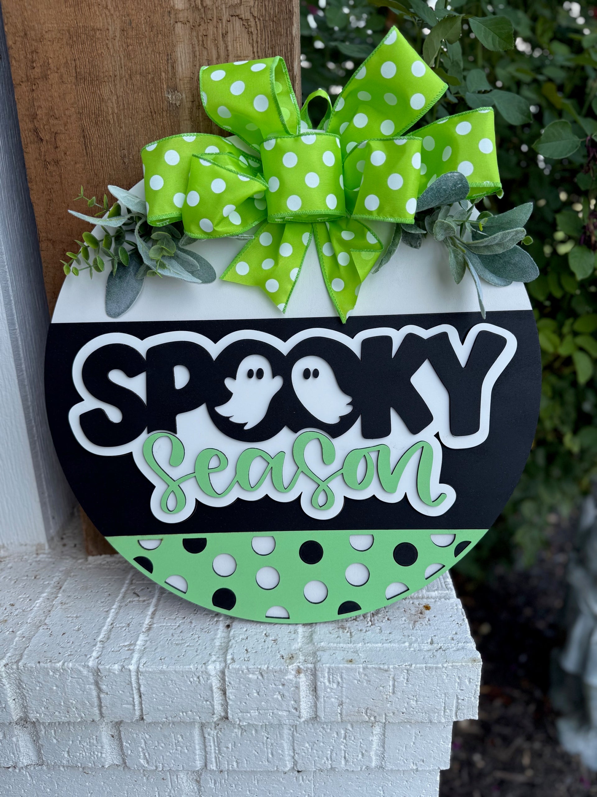 A round sign decorated with a green polka dot bow and greenery, featuring the words “Spooky Season” with ghost illustrations, in black, white, and green colors with polka dot accents, displayed outdoors.