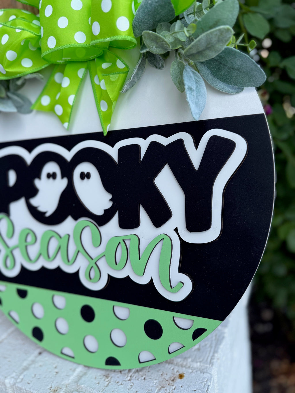 A decorative sign featuring the phrase Spooky Season with two cartoon ghost icons, green and white polka dots, and a green ribbon with white polka dots, surrounded by artificial leaves.