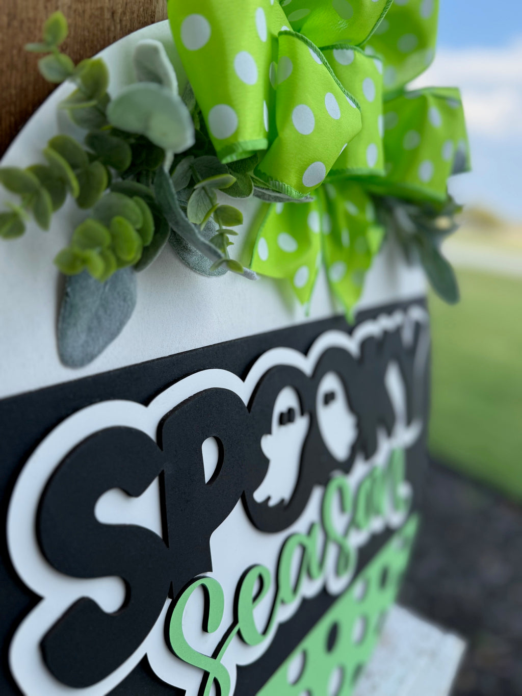 A close-up of a sign reading Spooky Season with ghost graphics, decorated with a green polka-dot bow and artificial greenery. The background is slightly blurred, showing an outdoor scene.