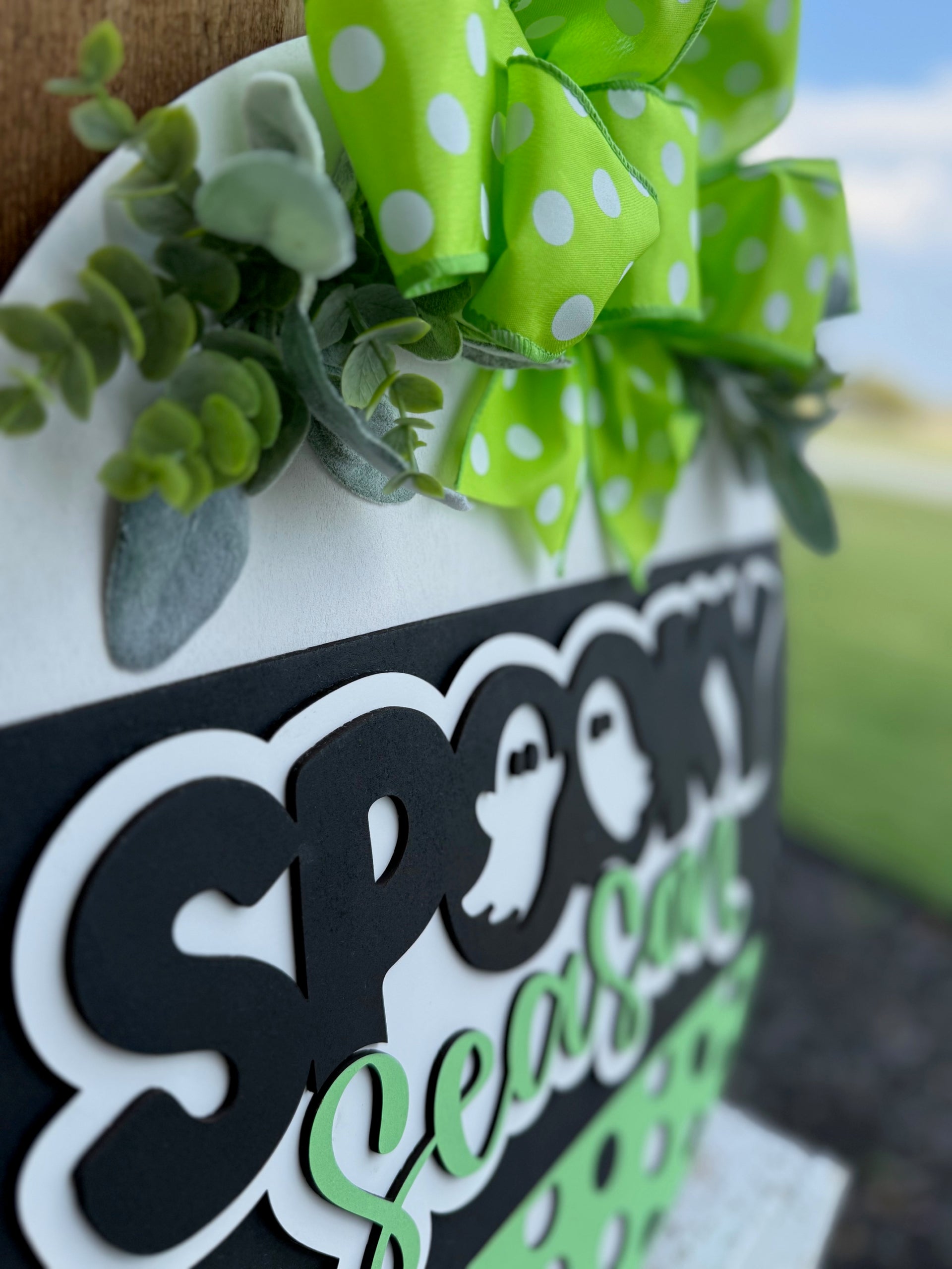 A close-up of a sign reading Spooky Season with ghost graphics, decorated with a green polka-dot bow and artificial greenery. The background is slightly blurred, showing an outdoor scene.