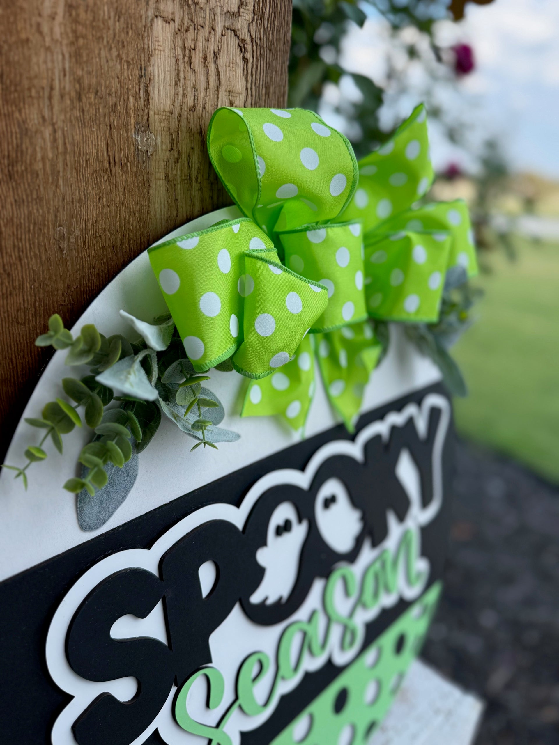 A close-up of a sign reading Spooky Season in black and green letters, decorated with a green polka dot bow and faux greenery, attached to a wooden post outdoors.