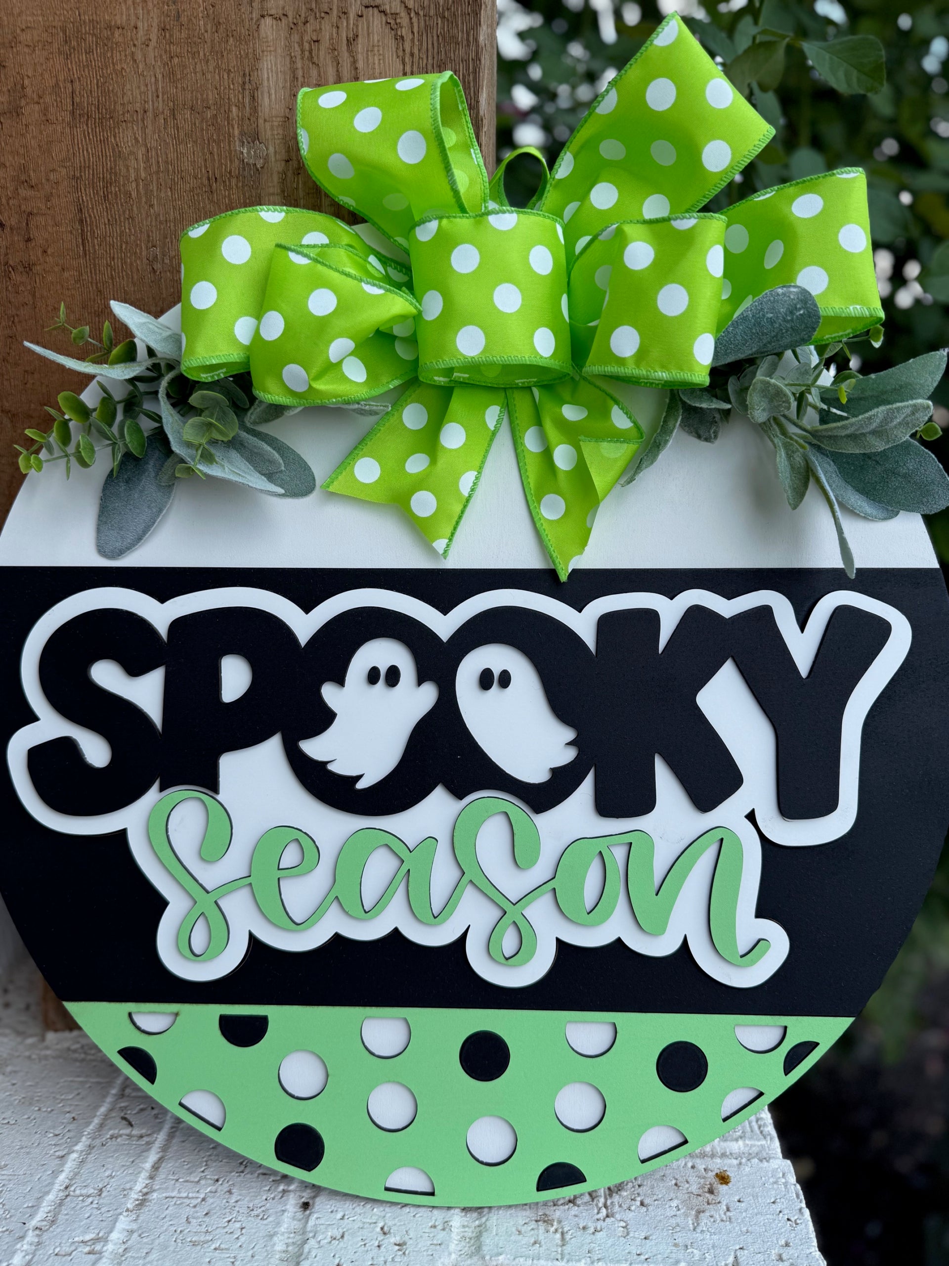 A round sign decorated with a green polka dot bow, faux greenery, and the words Spooky Season in black and green letters, with ghost illustrations for the Os, and black and green polka dots on the bottom.