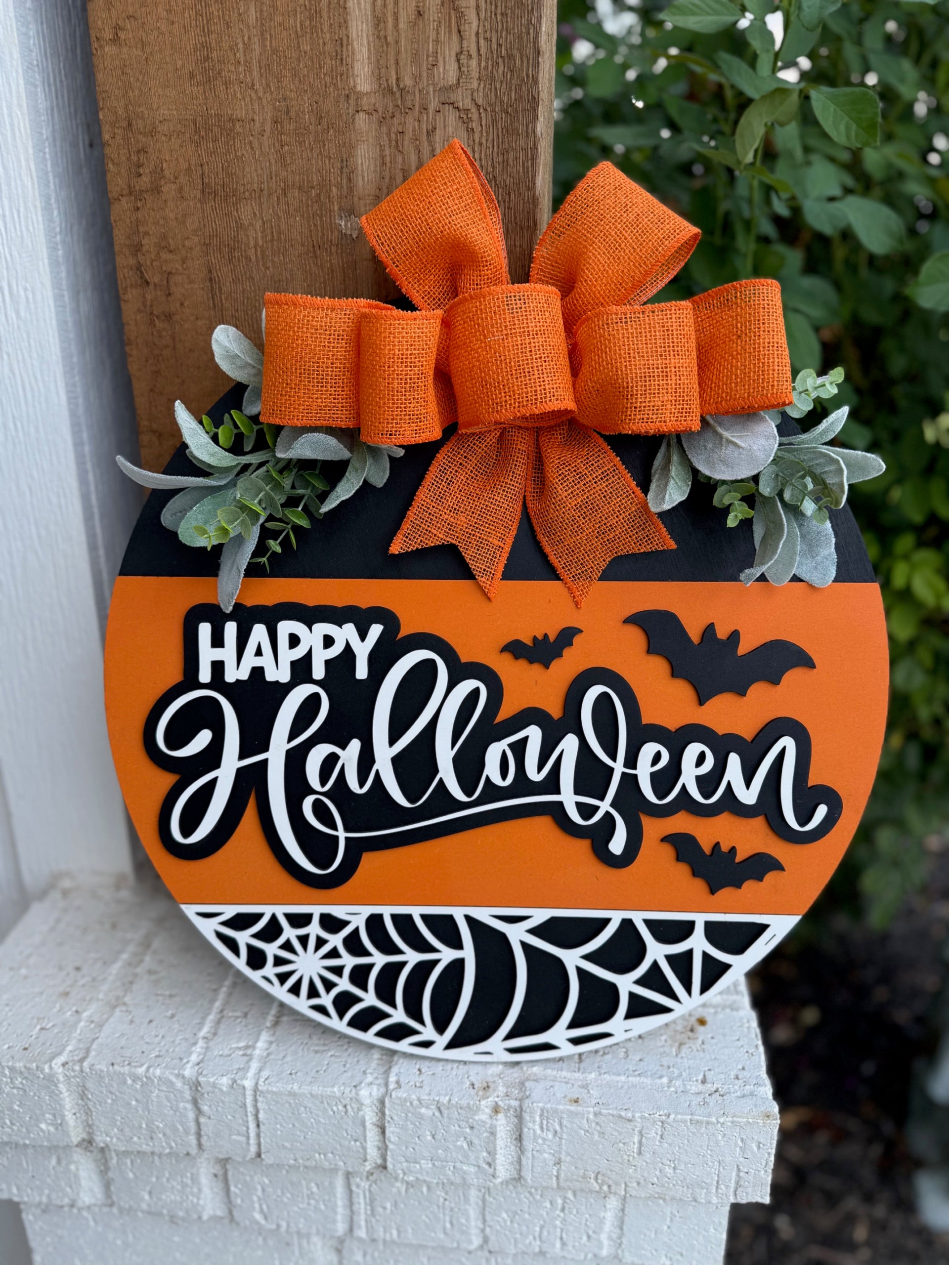 A round, orange-and-black Halloween sign with Happy Halloween written in white, decorated with bats, a spiderweb, greenery, and a large orange burlap bow at the top, hanging on a porch.