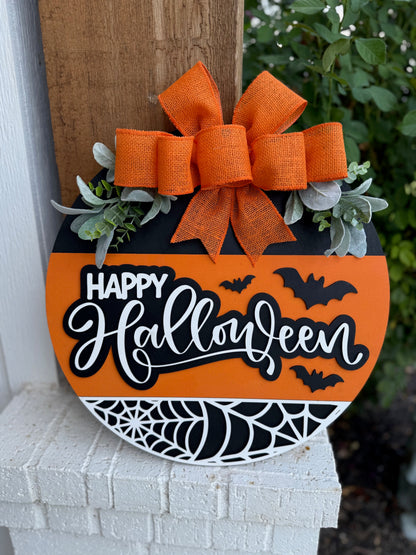 A round, orange-and-black Halloween sign with Happy Halloween written in white, decorated with bats, a spiderweb, greenery, and a large orange burlap bow at the top, hanging on a porch.